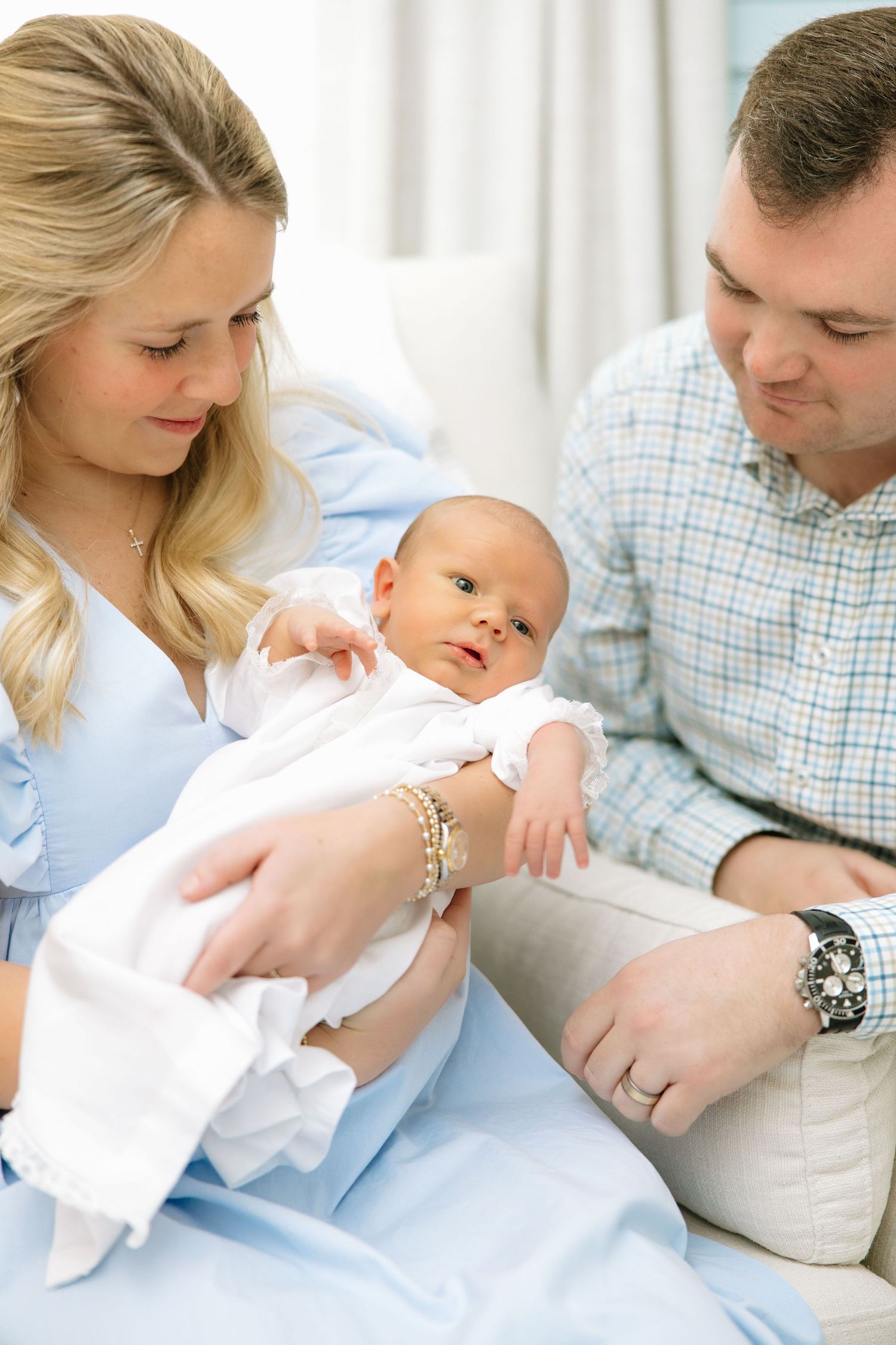 A young couple in blue and plaid attire lovingly gazes at their newborn baby wrapped in white.