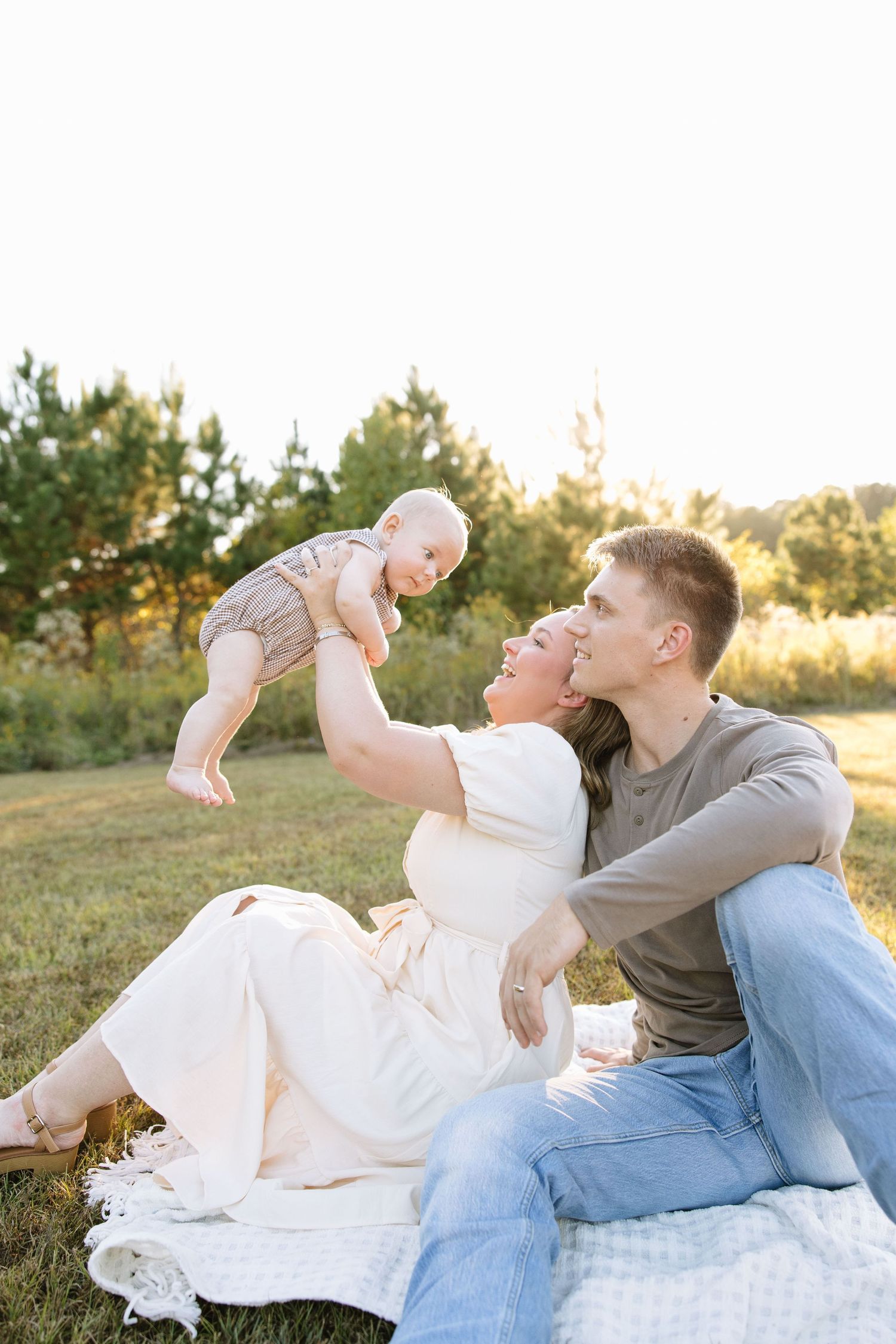 Happy young family sitting outdoors on blanket at sunset, mother lifting baby while father looks on lovingly.