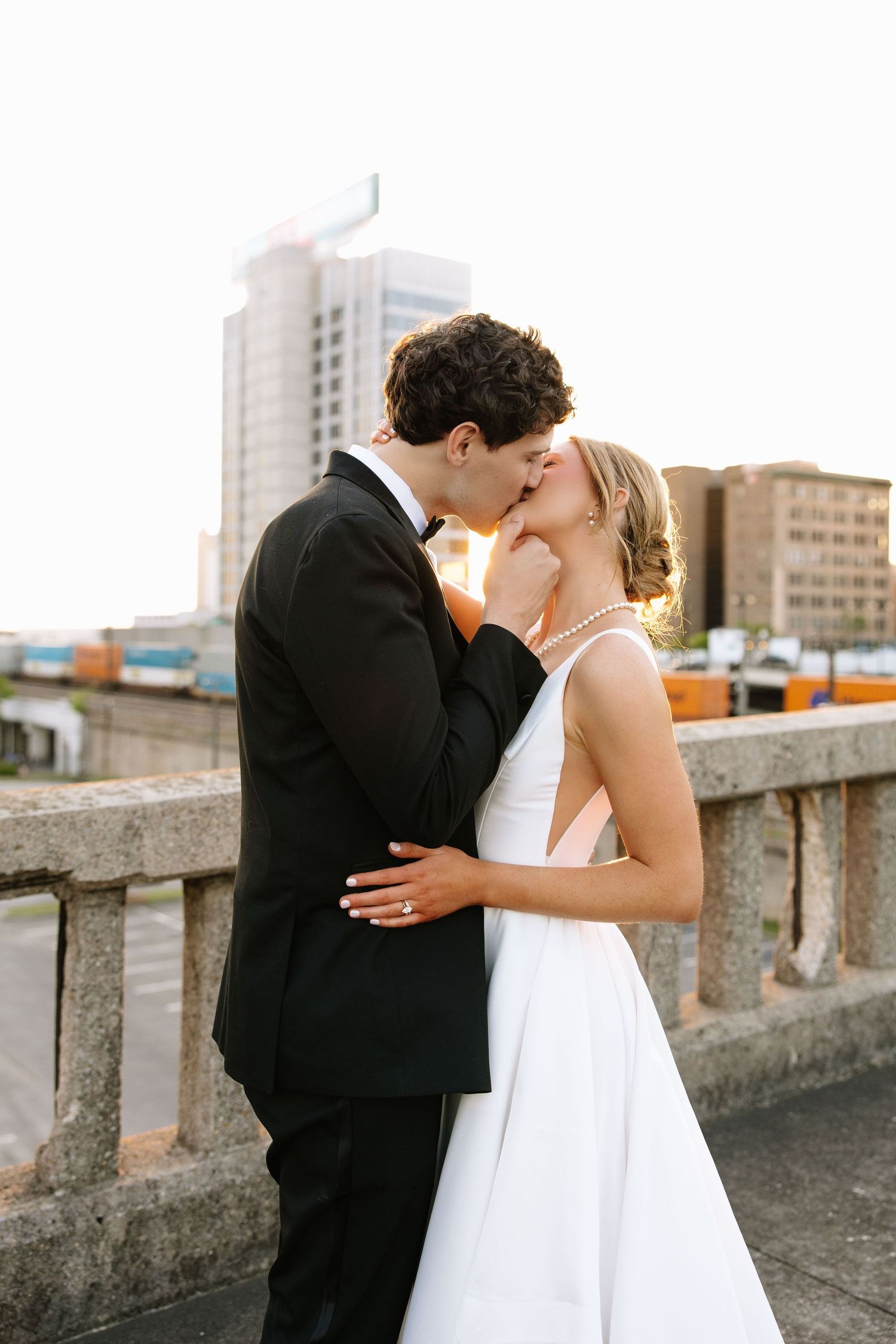 Wedding couple shares a tender kiss on a stone bridge with urban buildings visible behind them.