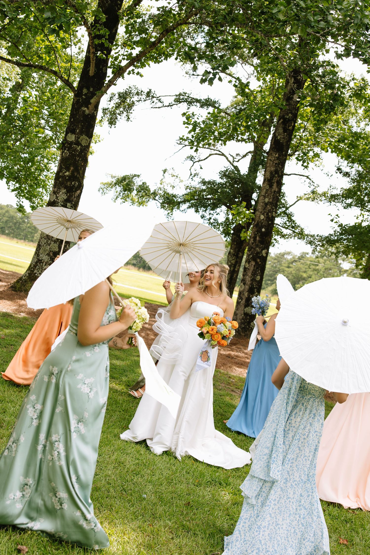 Bride in white gown with bridesmaids in colorful dresses holding white parasols outdoors on green lawn near trees.