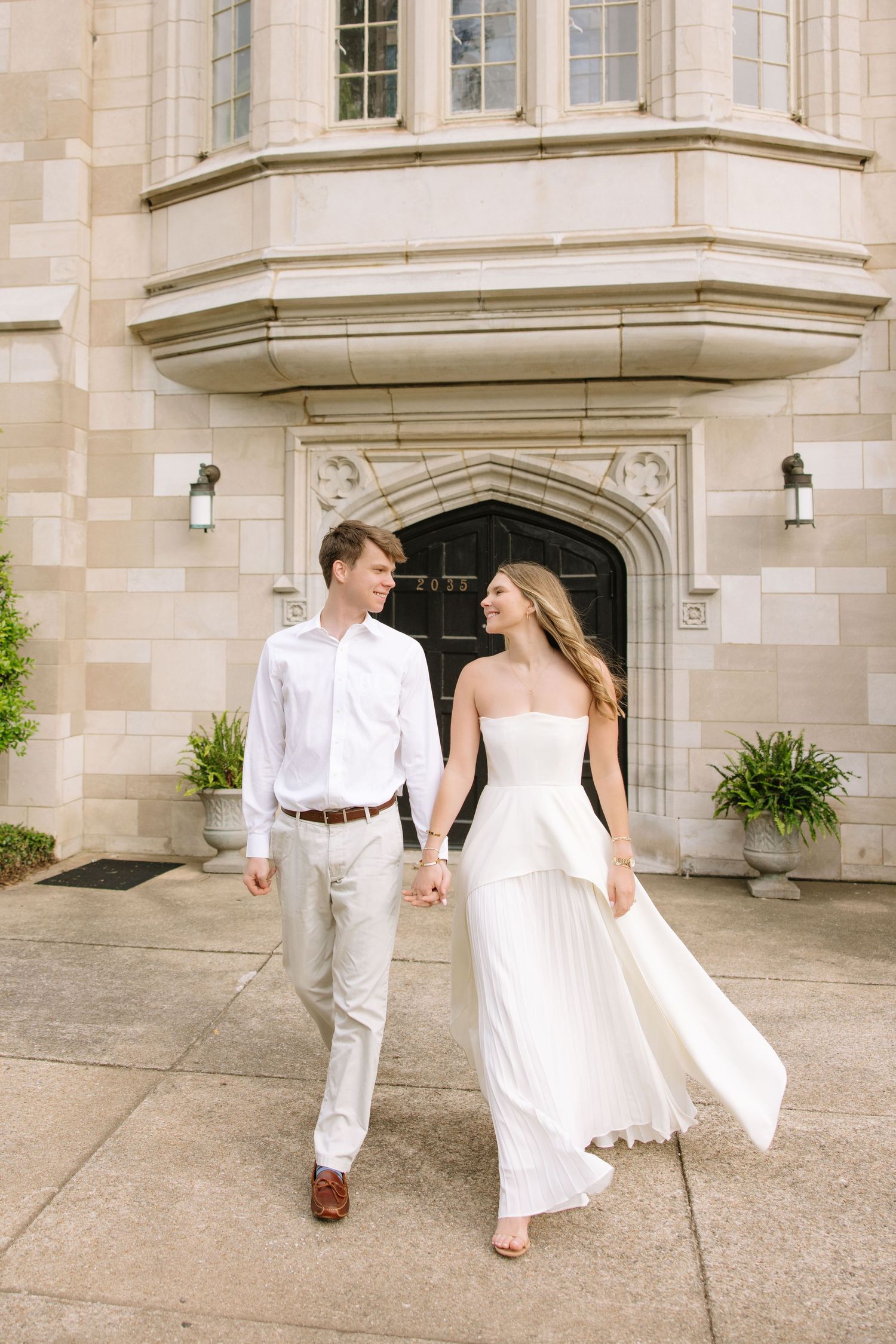 A couple in white attire holds hands walking in front of an elegant stone building on their wedding day.
