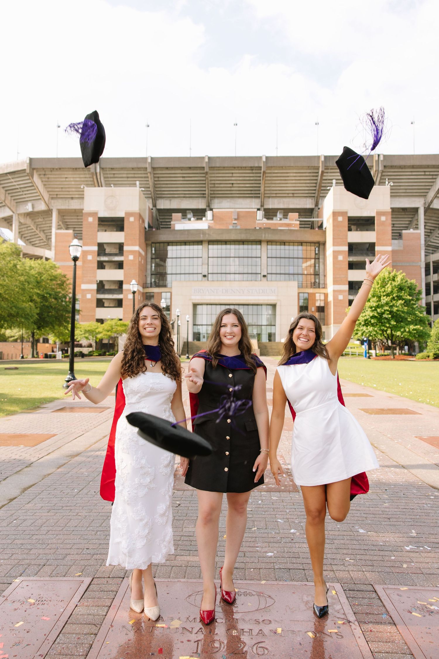 Three happy graduates toss their caps in front of a stadium, wearing white and black dresses with red capes.