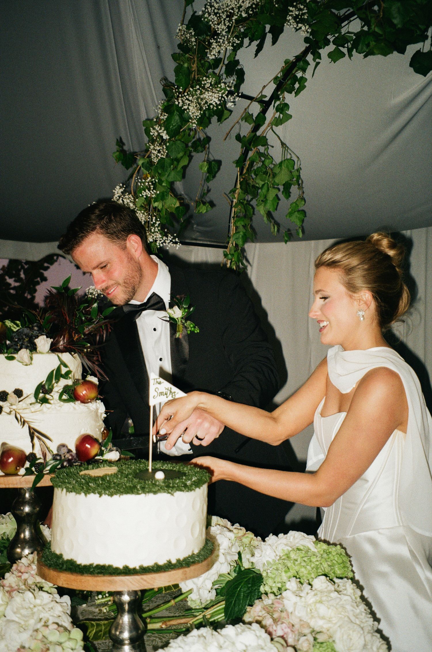 Bride and groom cutting a elegant white wedding cake decorated with moss, apples, and greenery under a tented reception.
