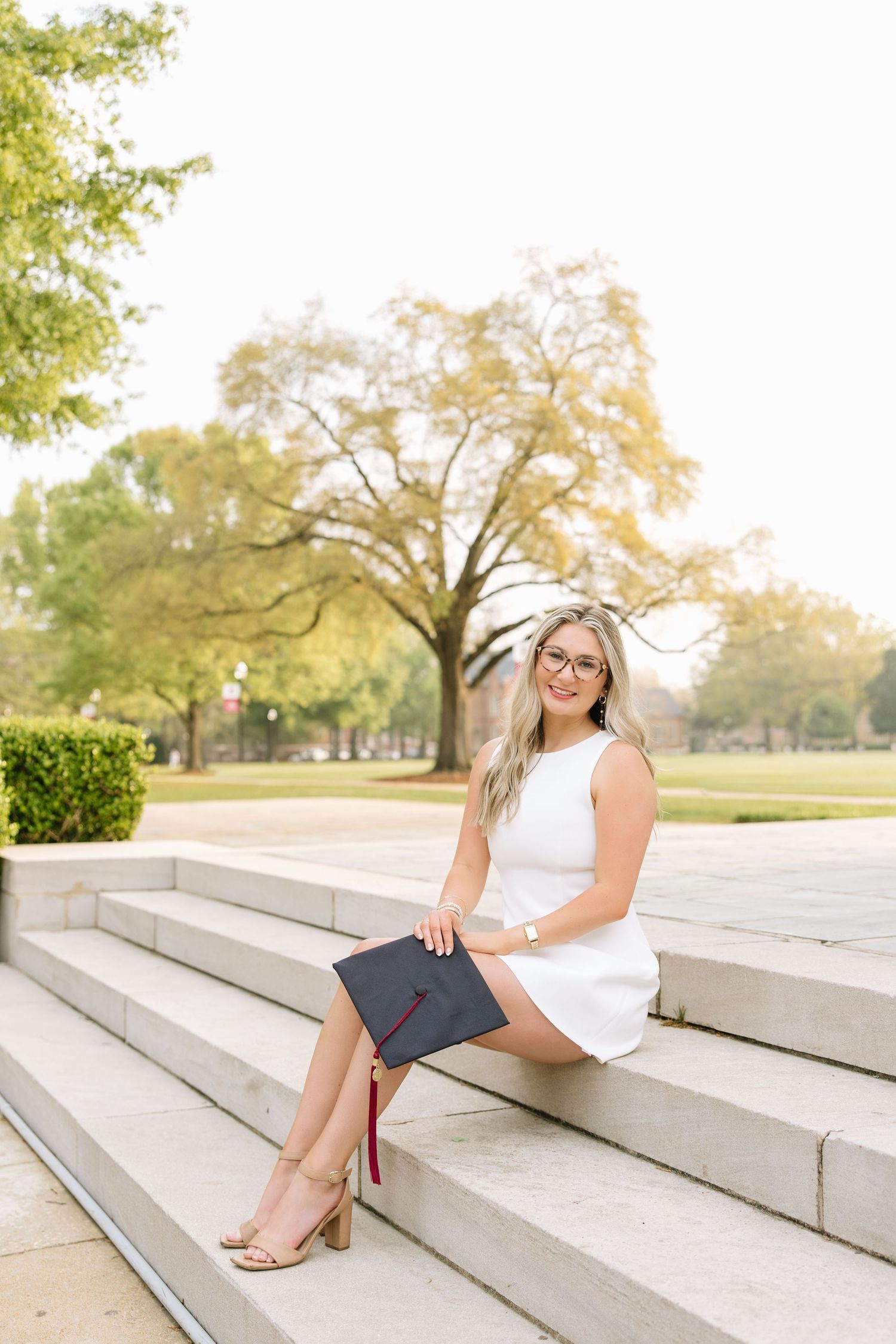 Young woman in white dress holding graduation cap sits on stone steps outdoors for senior portrait photo.