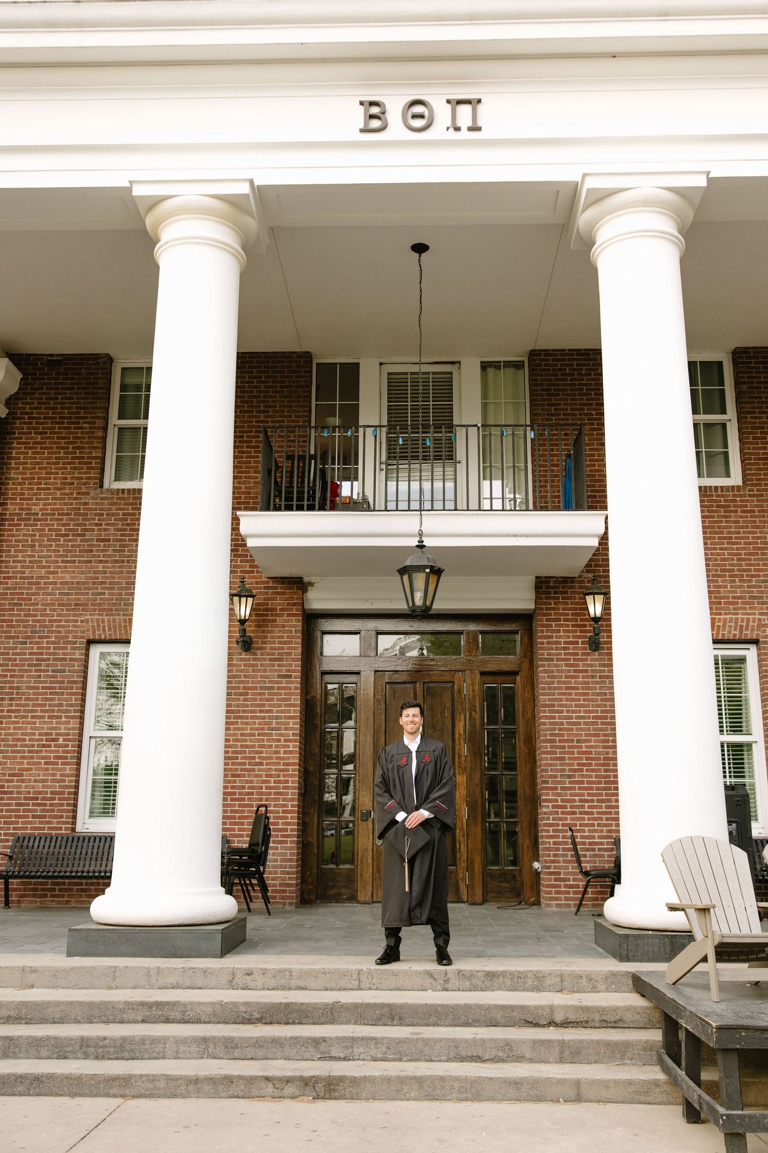 Man in graduation gown standing before Beta Theta Pi fraternity house with white columns and brick facade.