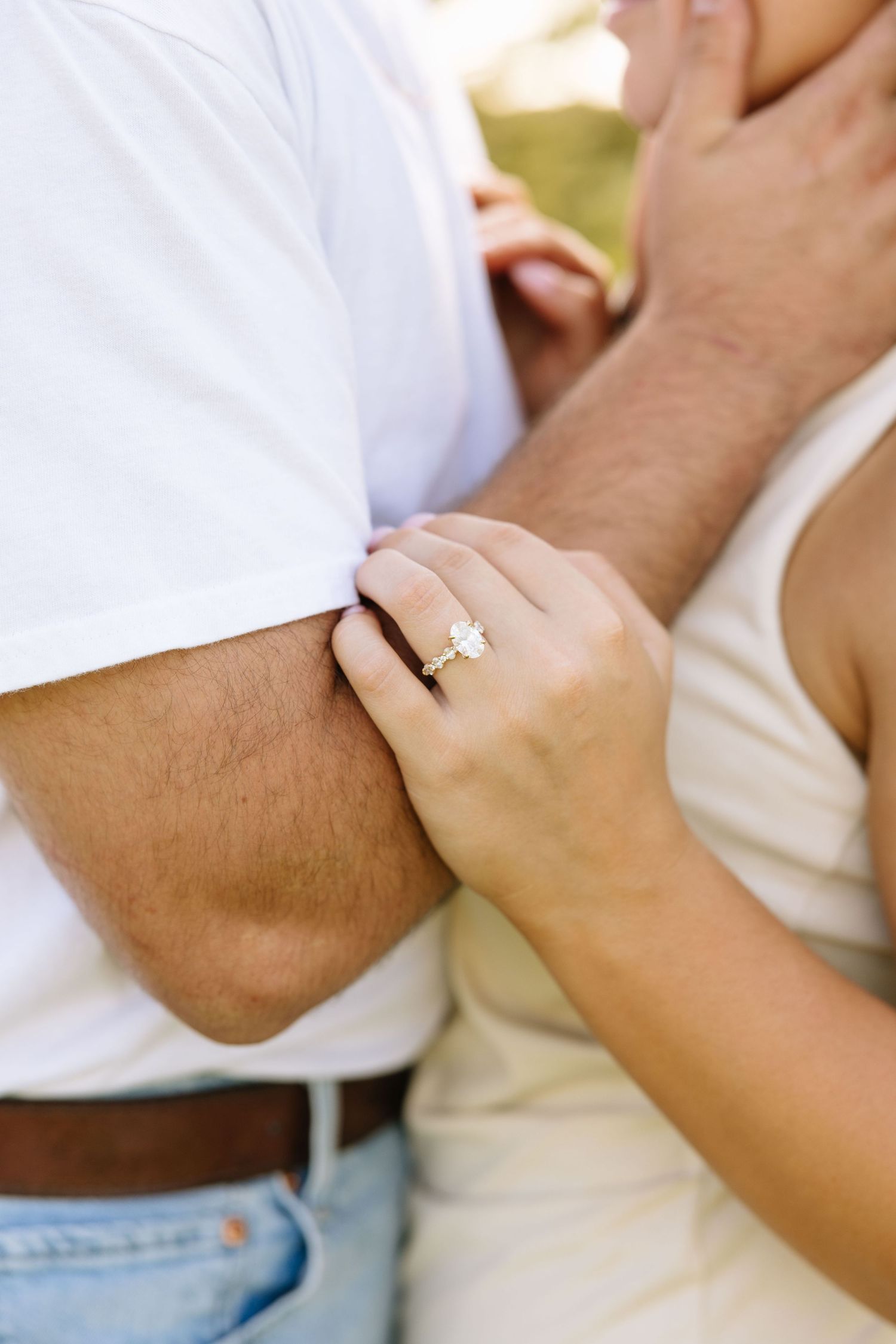 Close-up of a woman's hand with a diamond engagement ring resting on a man's arm while embracing outdoors.