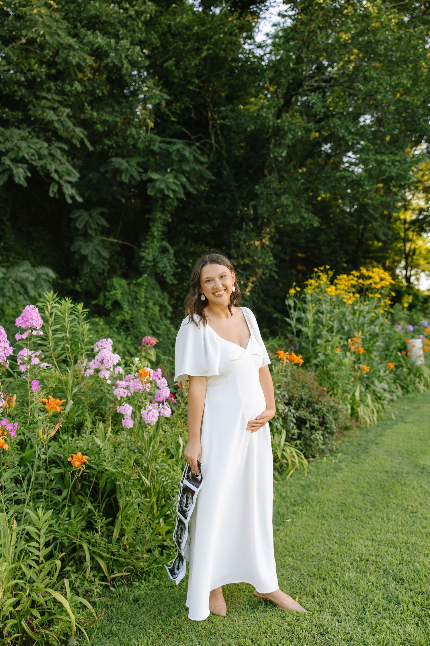 Smiling pregnant woman in white maxi dress stands barefoot in lush garden holding ultrasound images.