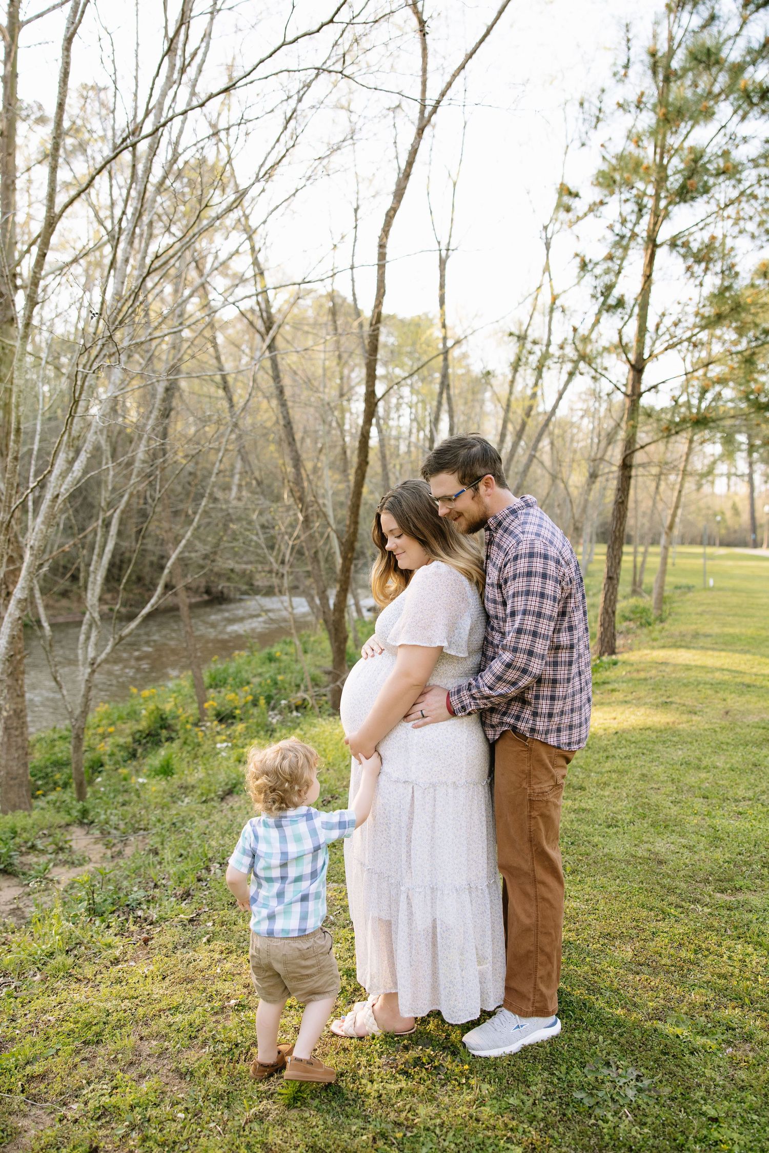 Pregnant woman in white dress with husband and toddler son posing outdoors near trees during maternity photo session.