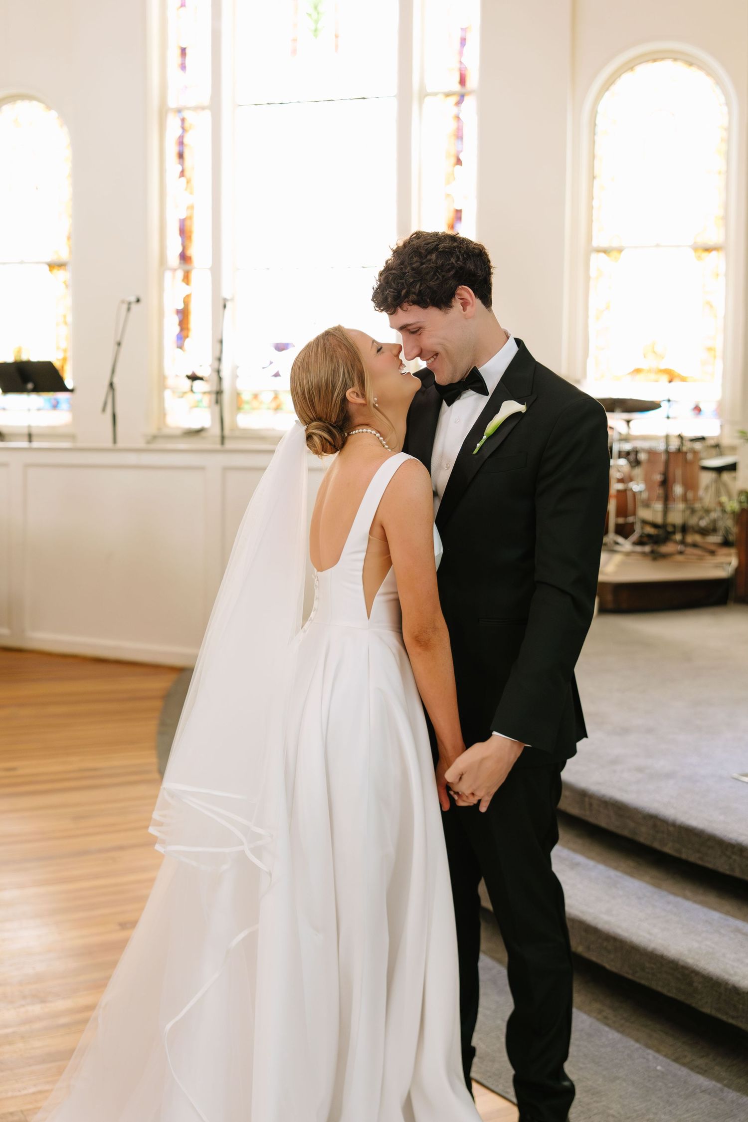 Bride and groom share a romantic kiss in a sunlit church, bride in elegant white gown with veil, groom in black tuxedo.