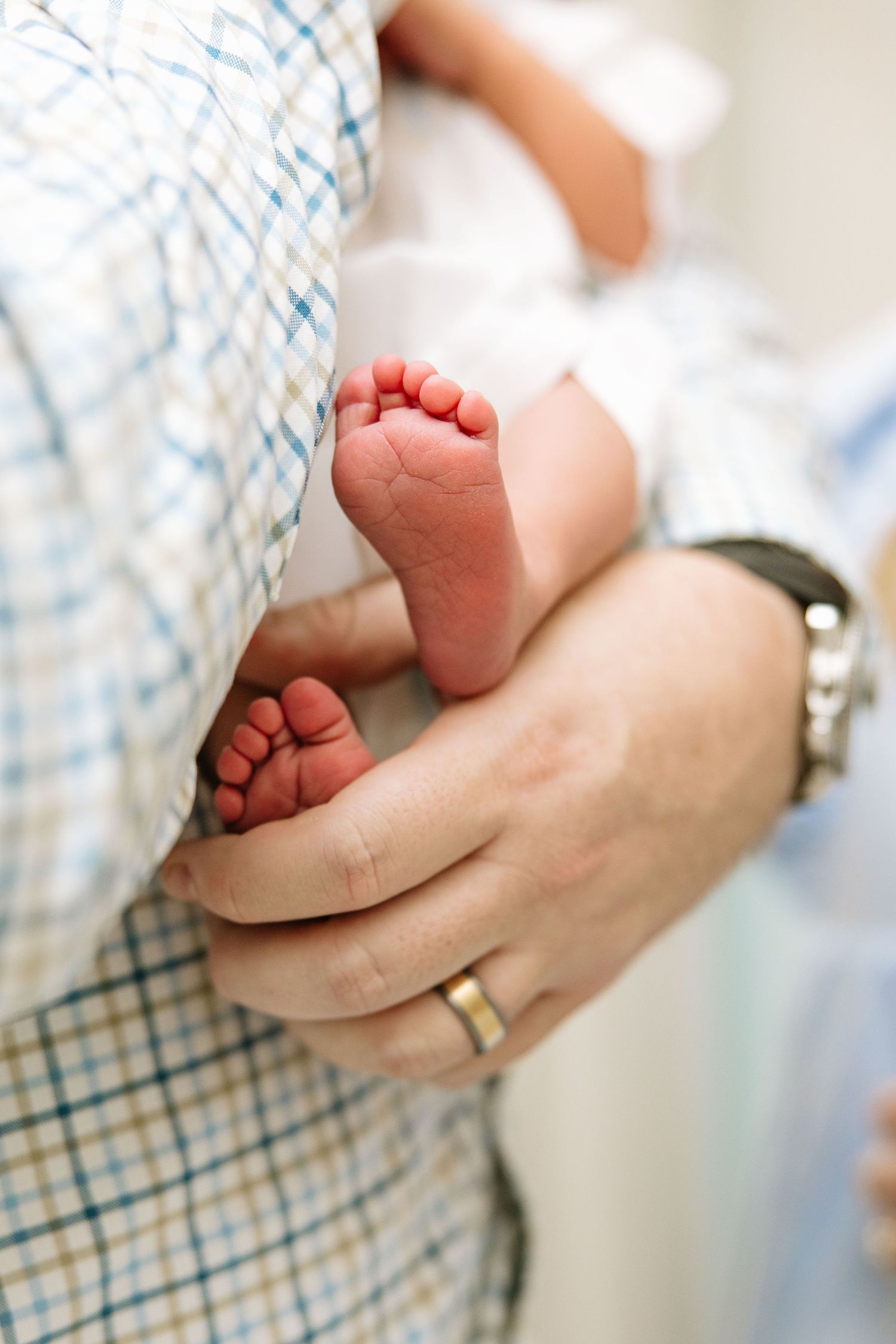 Close-up of a newborn baby's tiny feet cradled in a father's hands, wearing a wedding ring and watch.