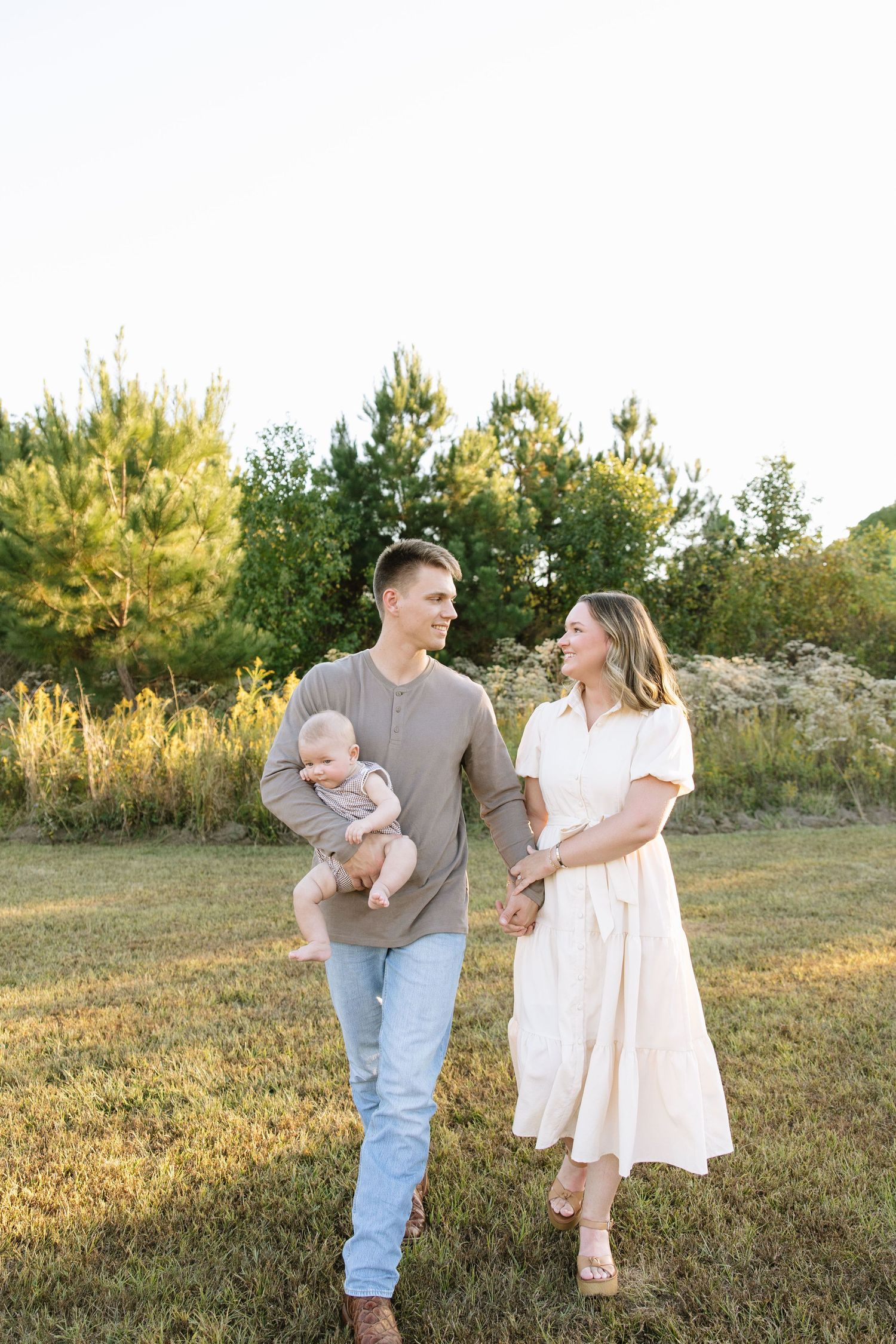 Young couple walking outdoors in golden hour light, man holding baby, woman in white dress, green trees in background.
