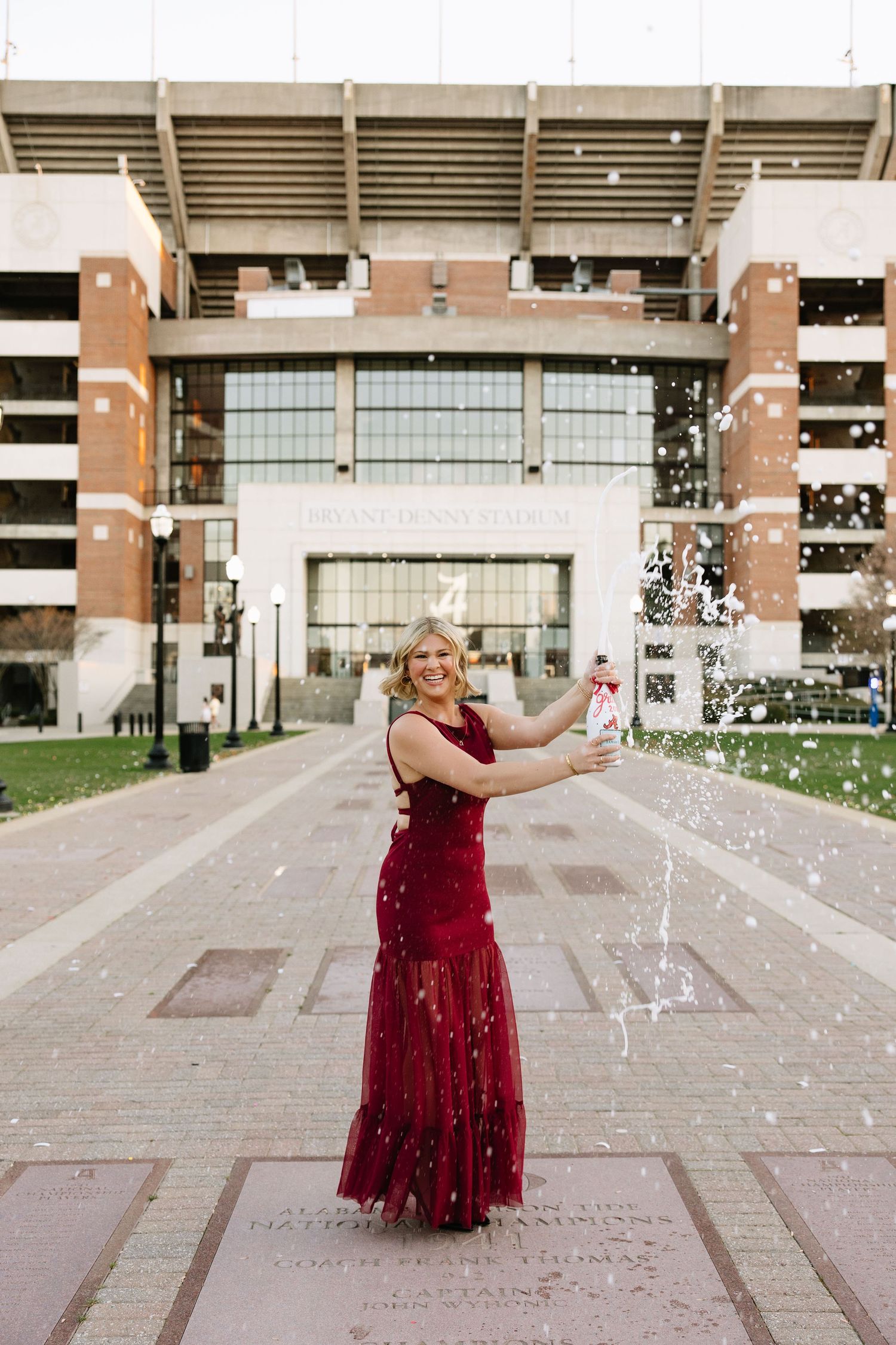 Woman in red gown celebrates with confetti outside Bryant-Denny Stadium at University of Alabama.