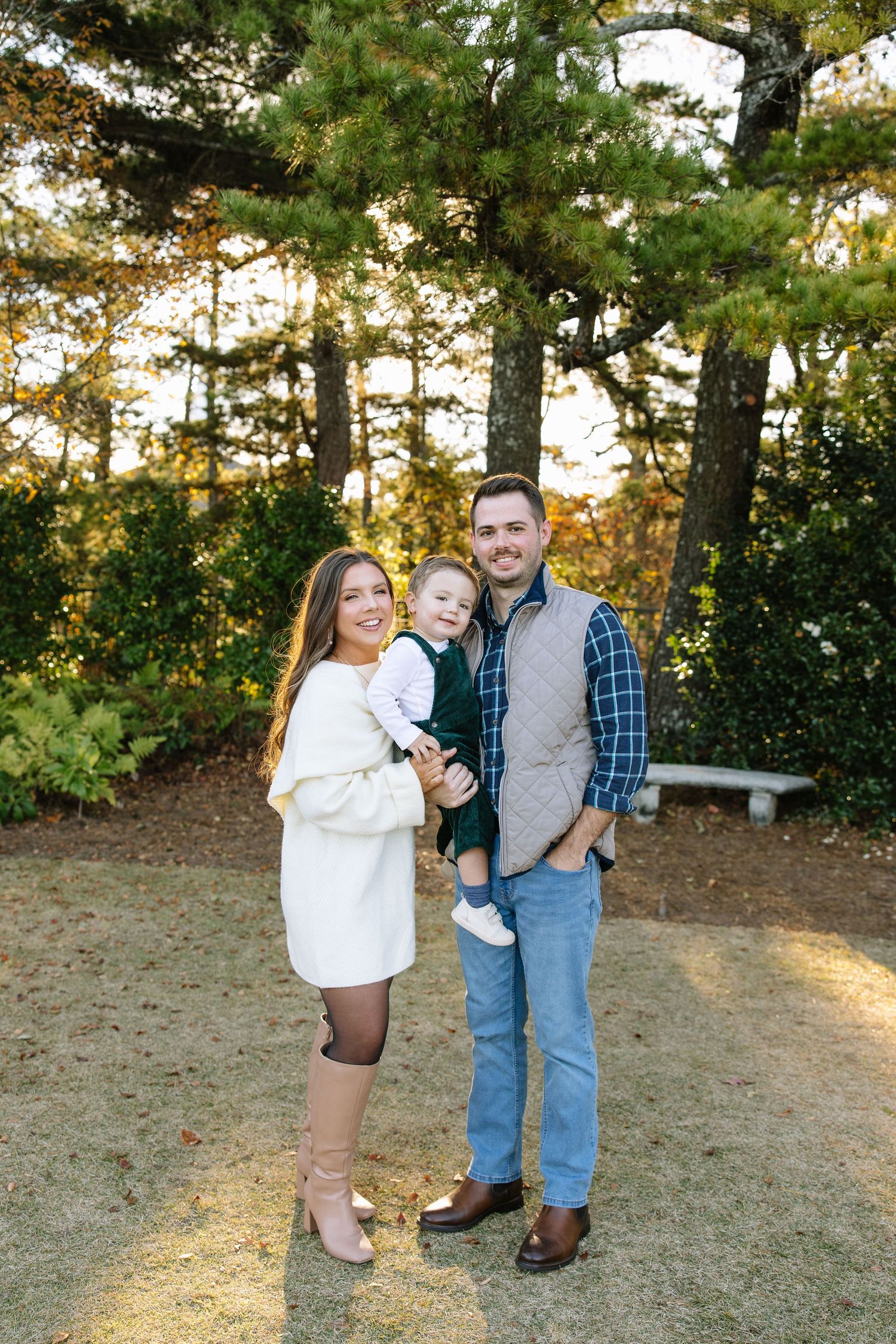 A young family of three poses outdoors, parents holding toddler, surrounded by autumn trees in golden light.