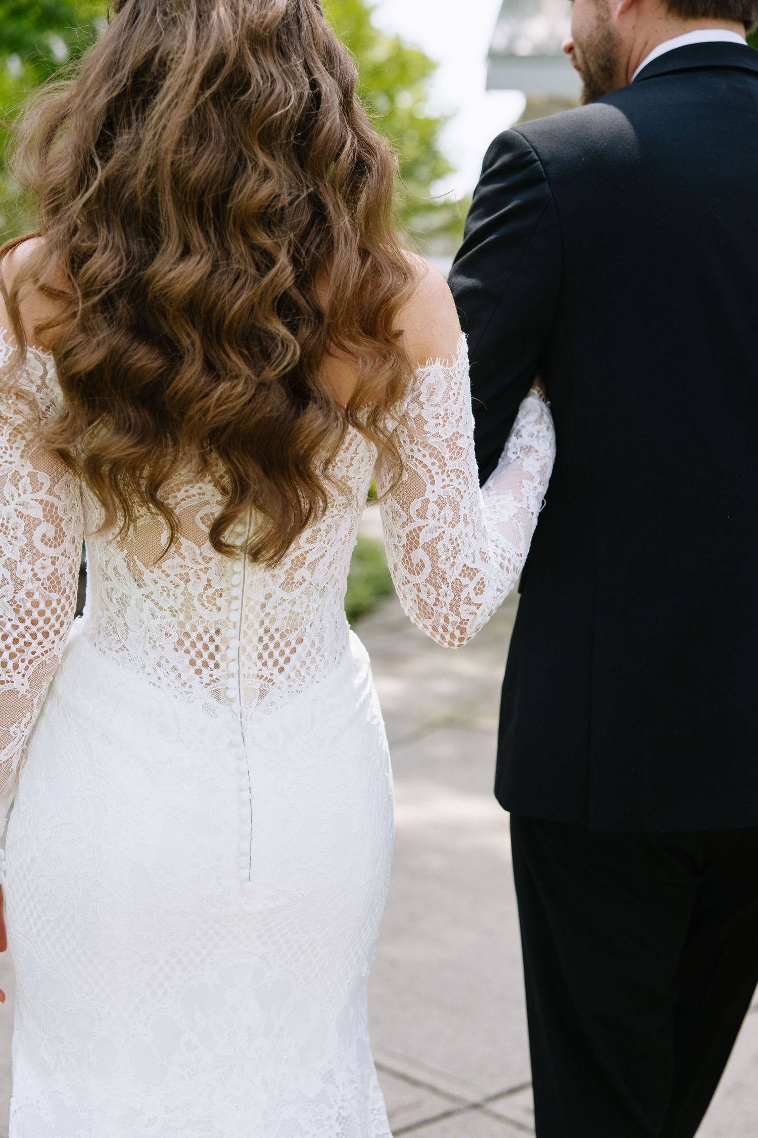 Close-up view of bride in lace long sleeve wedding dress with flowing curled hair standing next to groom in black suit.