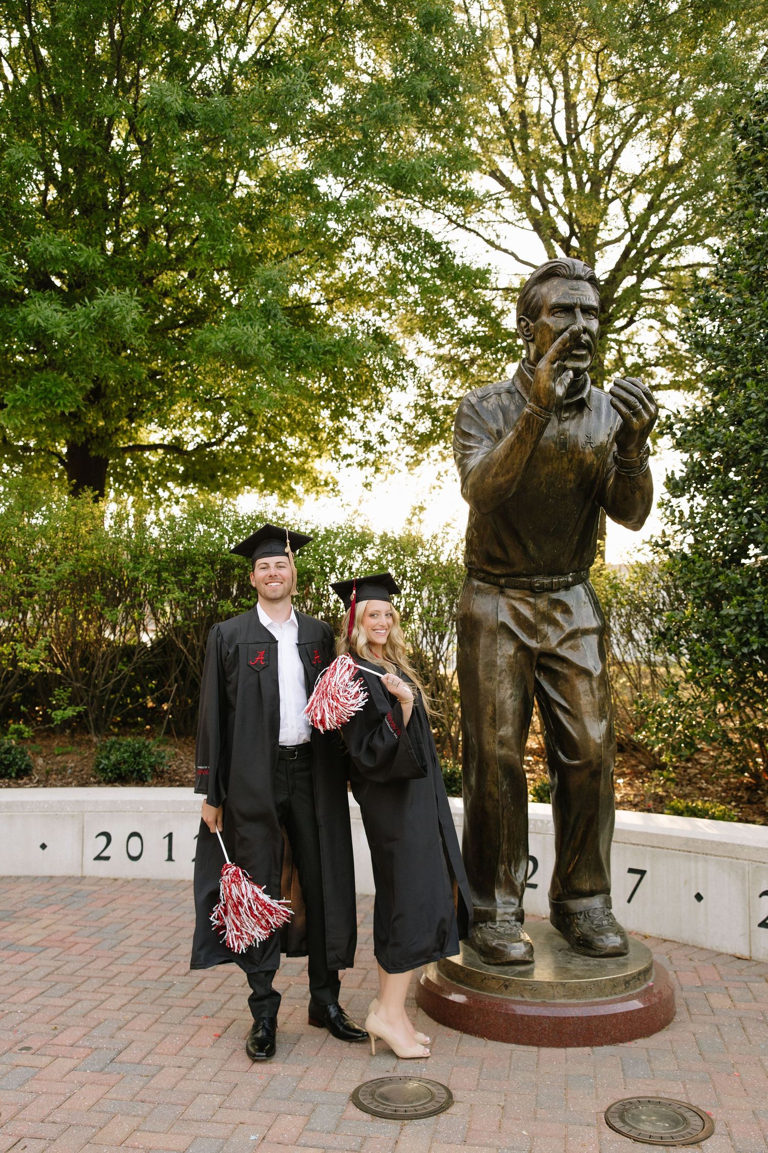 Two graduates in black caps and gowns pose beside a bronze coach statue on a university campus.