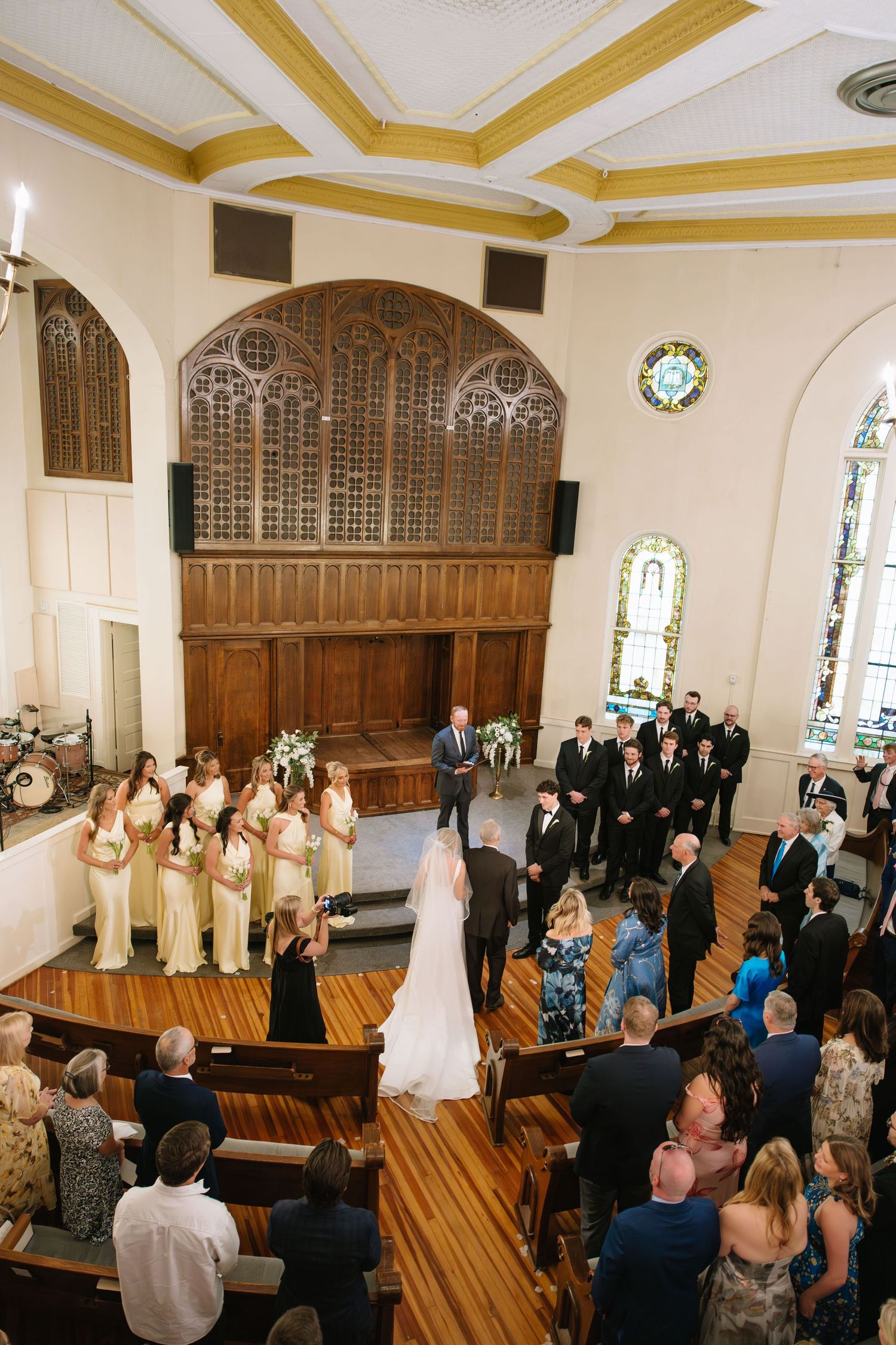 Aerial view of traditional church wedding ceremony with bridesmaids in yellow gowns and groomsmen in black suits.