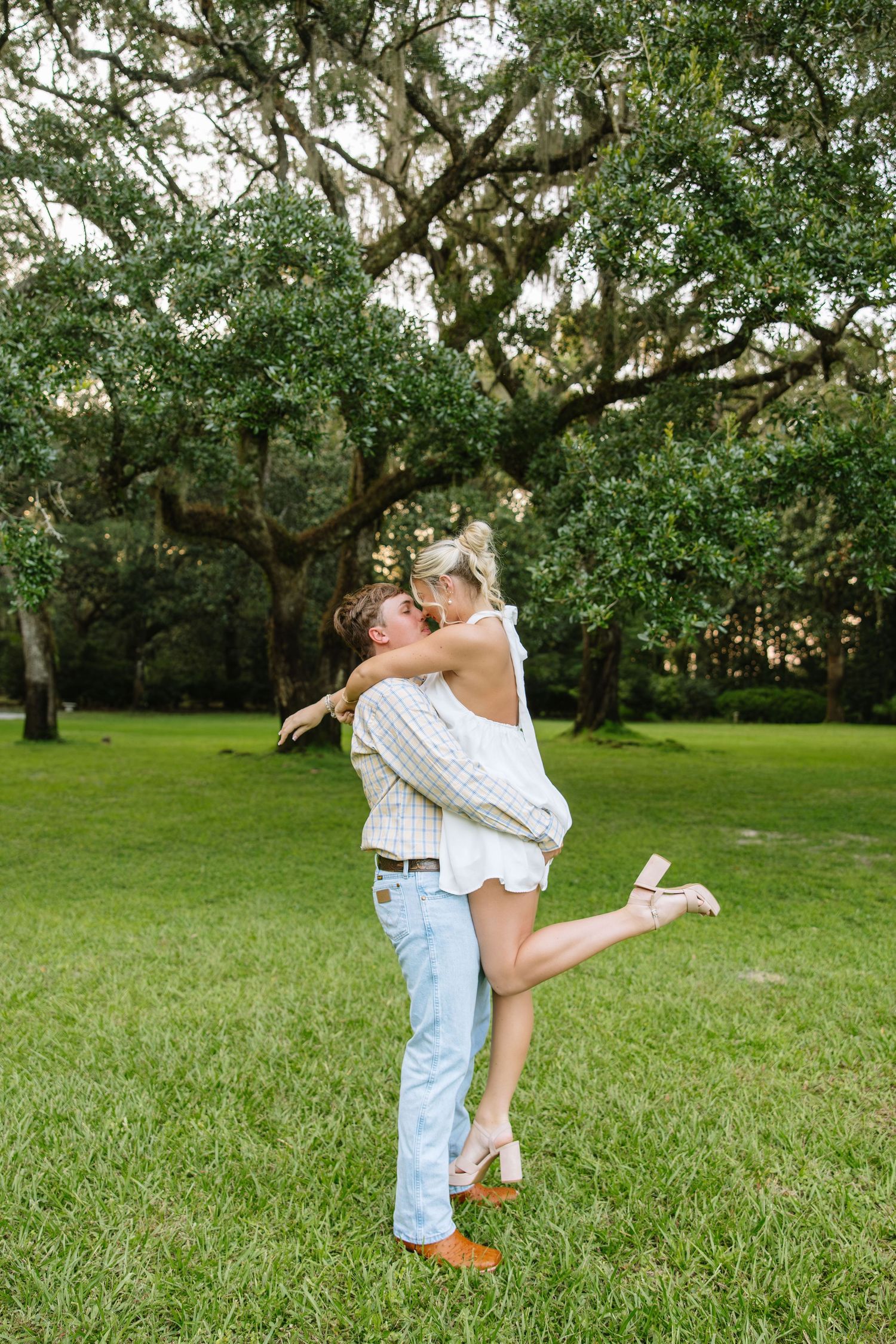 Couple sharing a romantic kiss outdoors, man lifting woman in white dress on lush green lawn beneath sprawling oak trees.