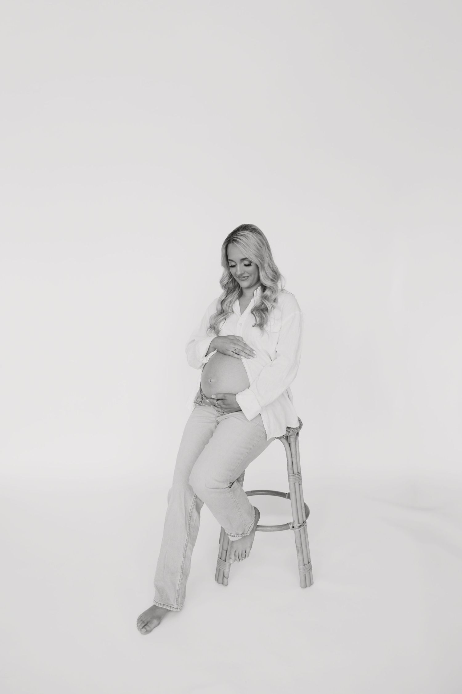 Black and white maternity photo of a pregnant woman in a white shirt and jeans sitting on a stool.