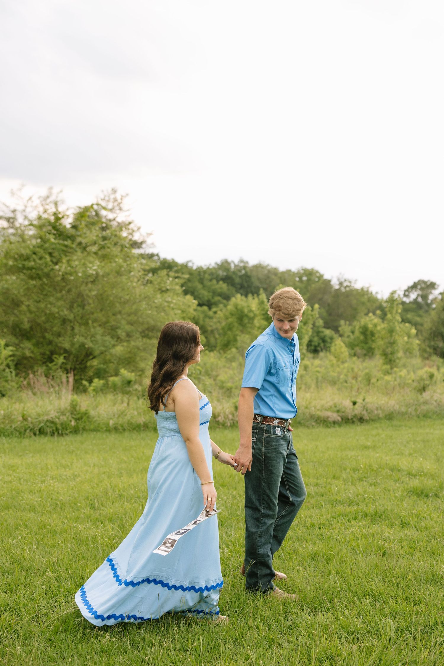 A young couple holds hands in a green field, the girl wearing a flowing blue dress and the boy in a blue shirt and dark pants.
