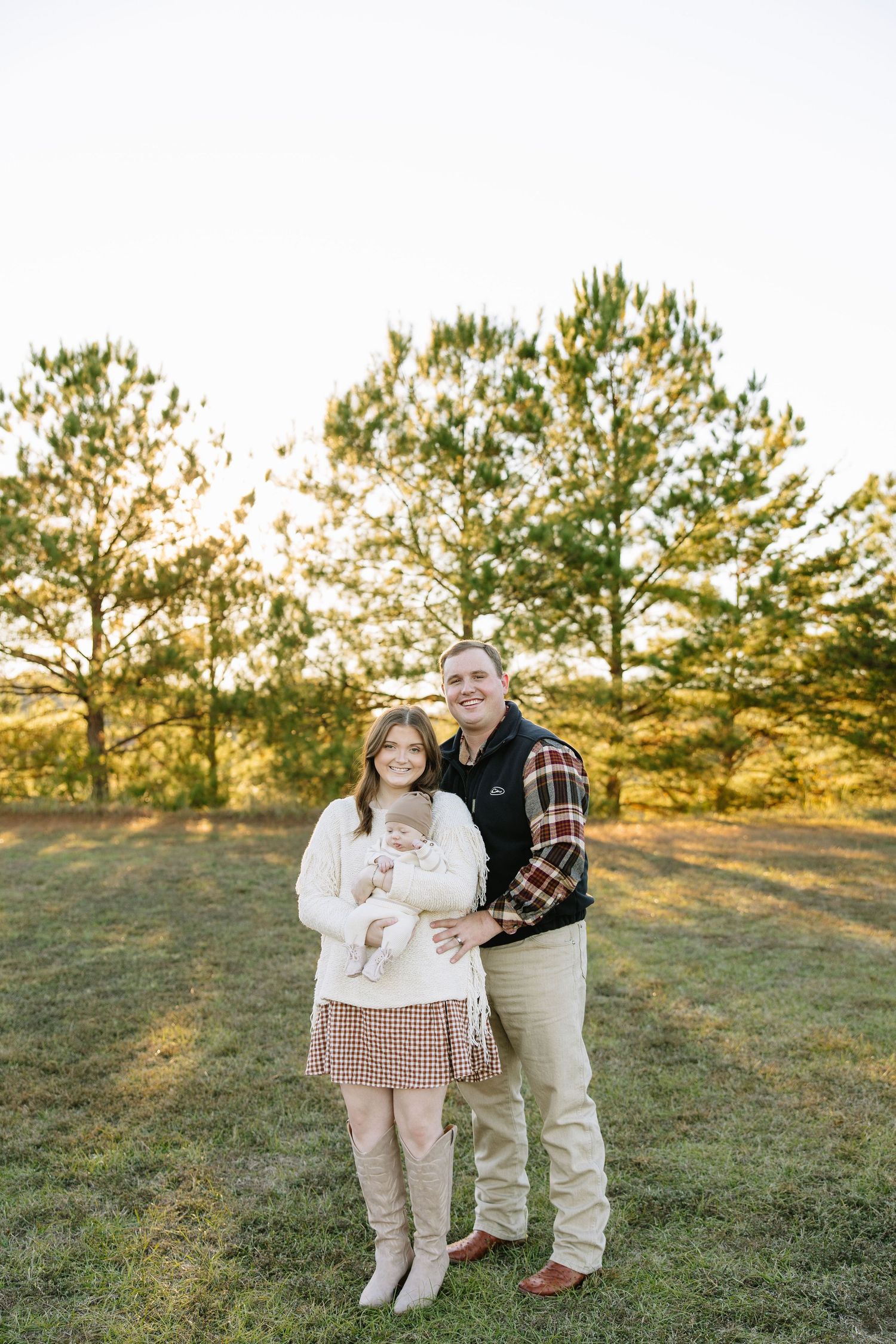 A young family poses outdoors at golden hour, mother holding infant, father beside her, pine trees in background.