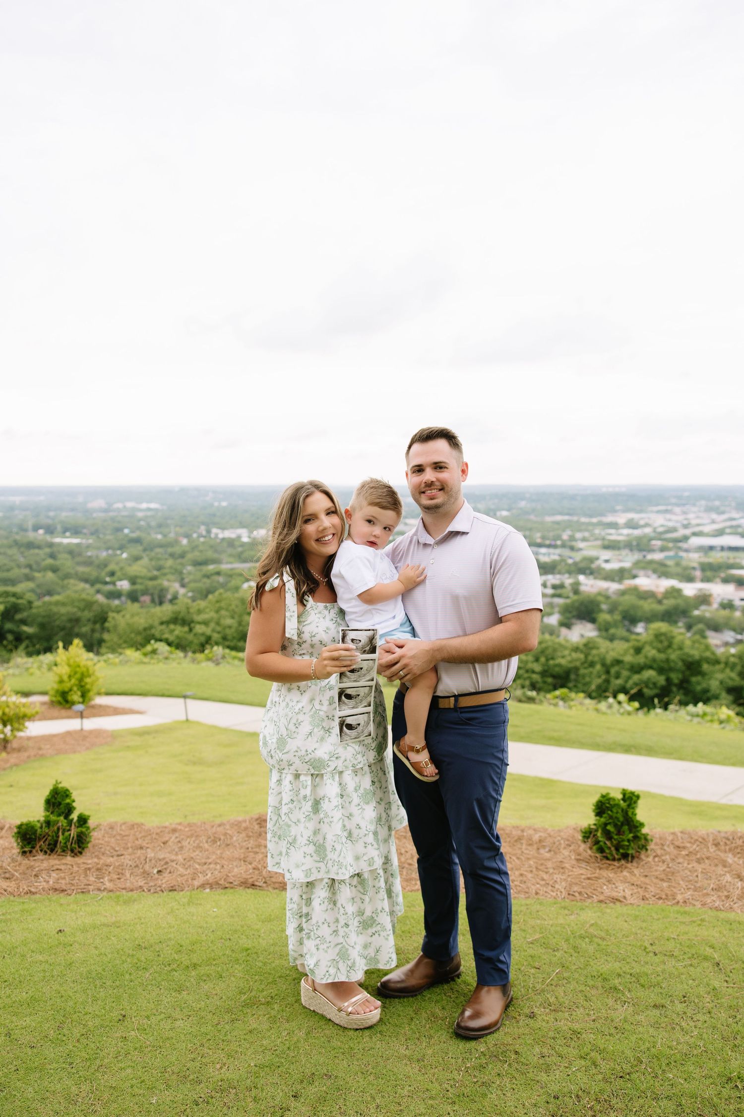 Happy family of three posing outdoors on a green hillside, woman in floral dress holding ultrasound photos.