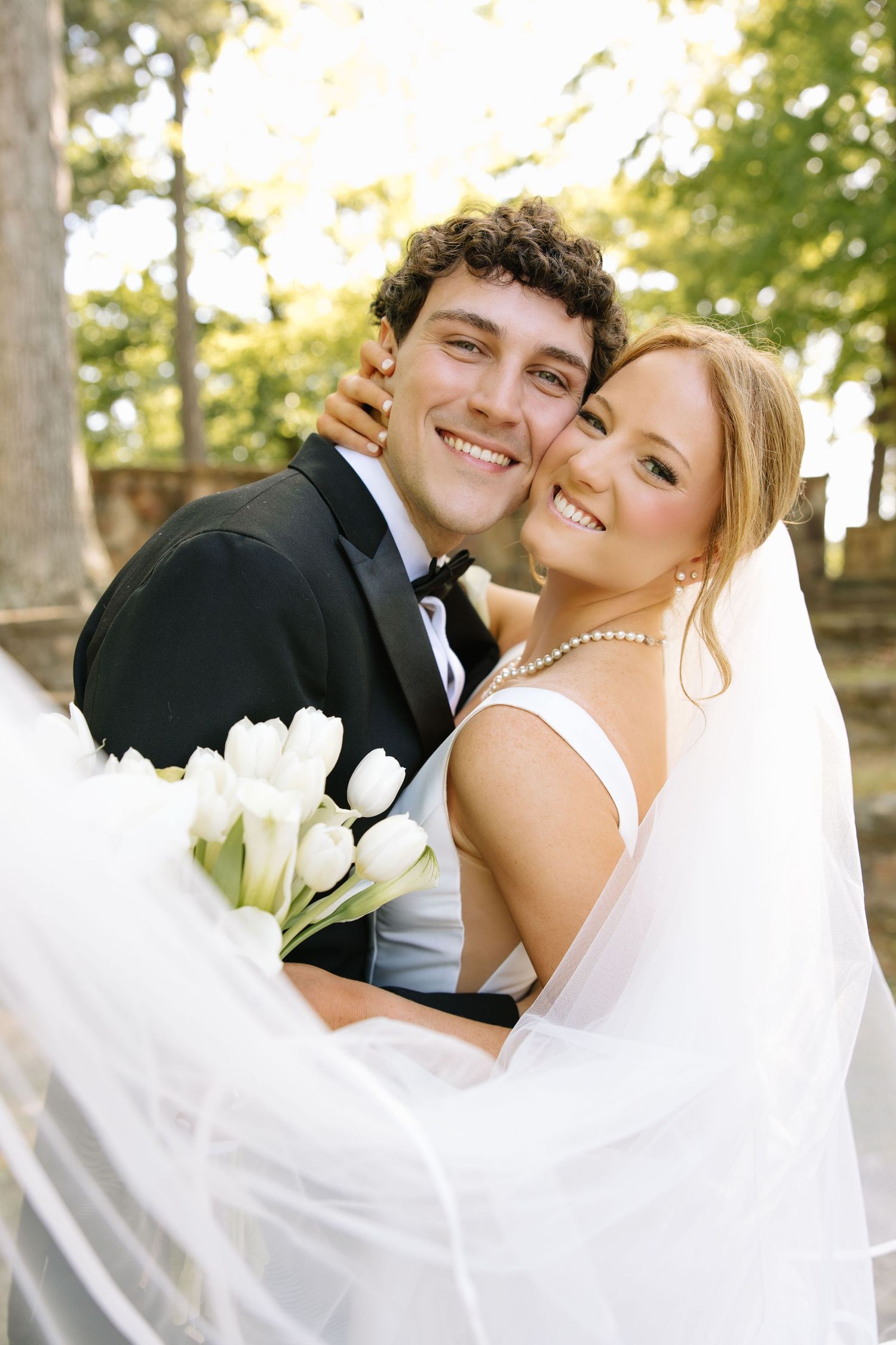 Smiling bride and groom embrace outdoors, bride holding white tulips bouquet and wearing veil and pearl necklace.
