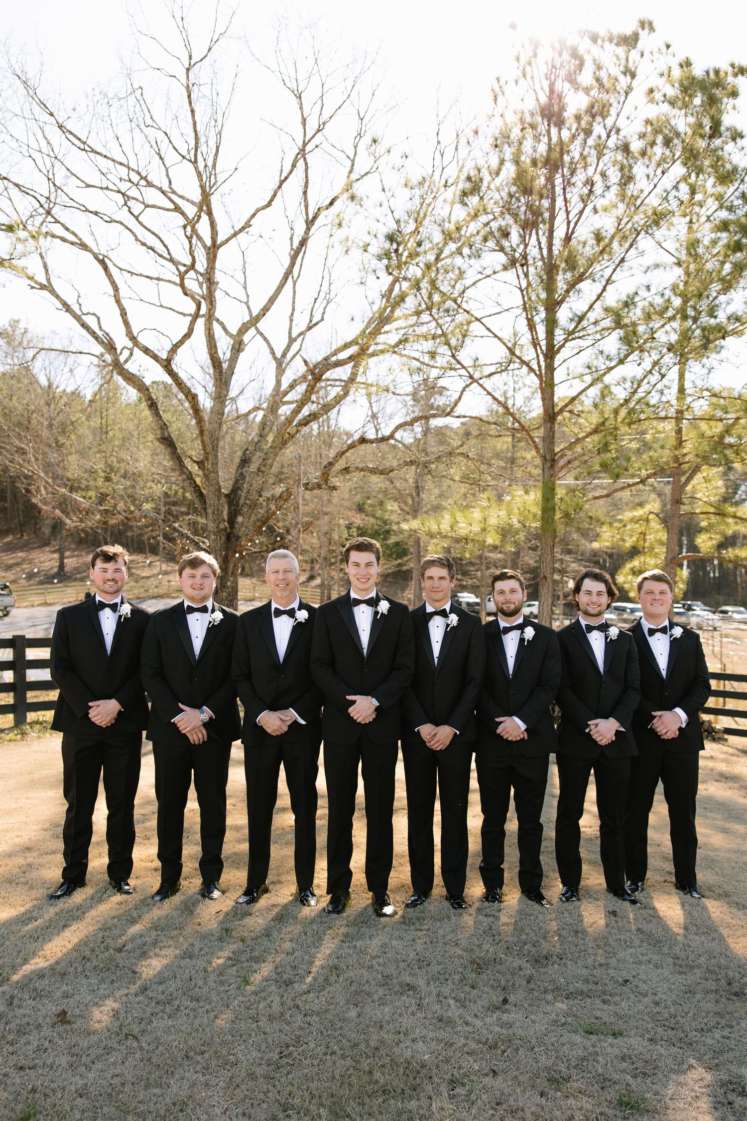 Groomsmen in black tuxedos stand in a line outdoors during a wedding.