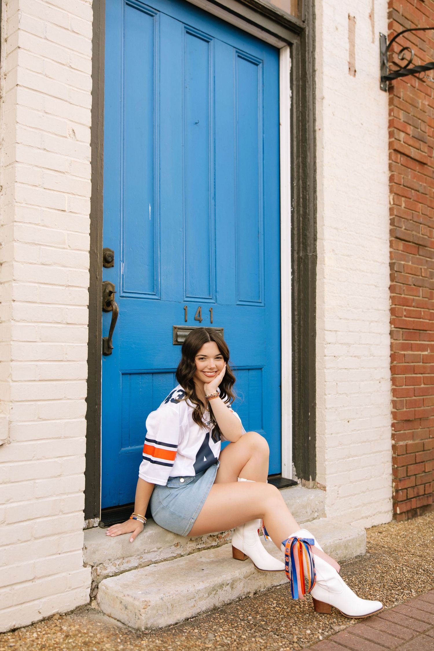 Young woman in white jersey and denim skirt sitting on steps in front of a bright blue door numbered 141.
