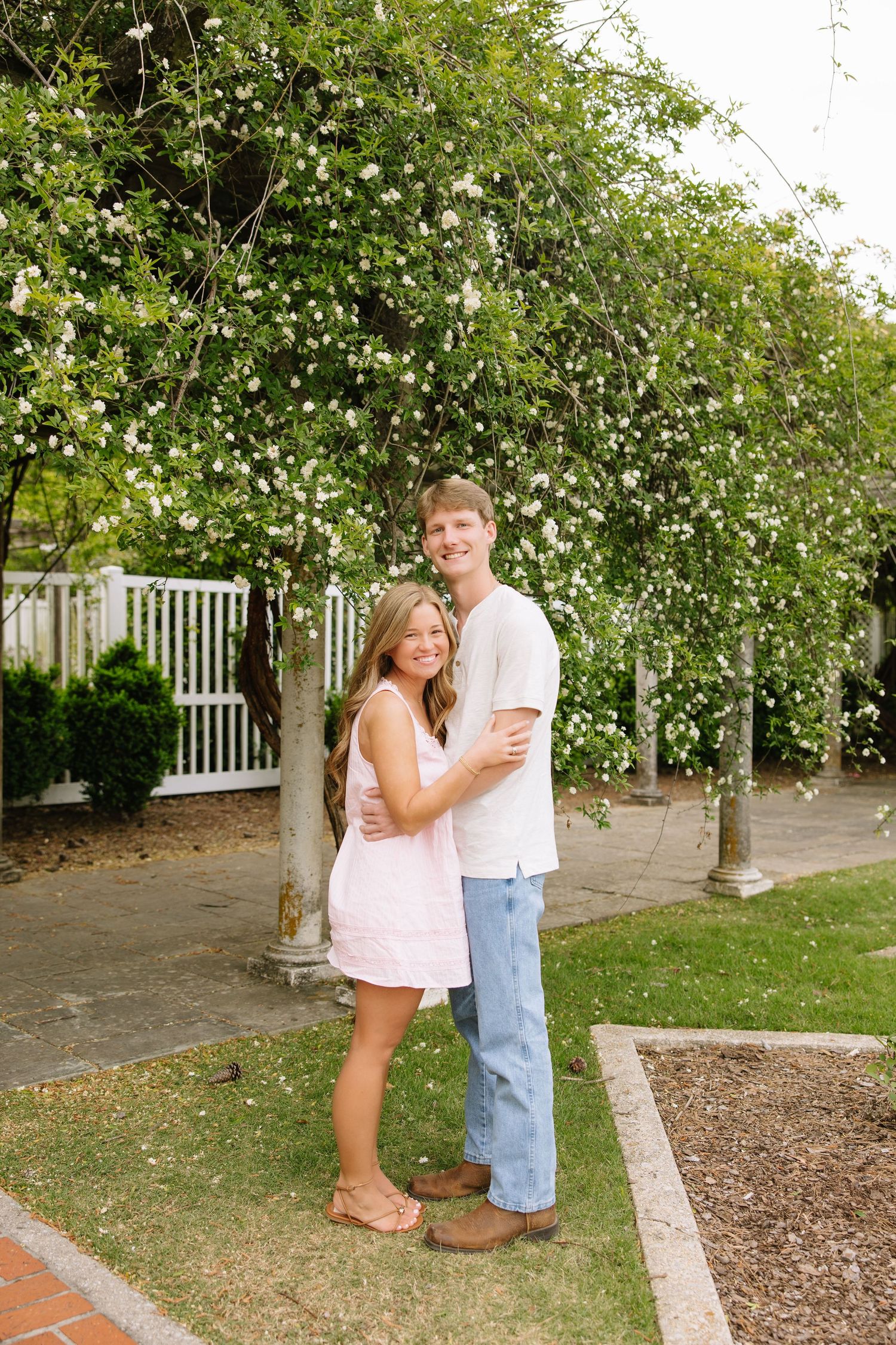 A smiling couple embraces outdoors near a flowering white rose trellis and white picket fence in a garden.