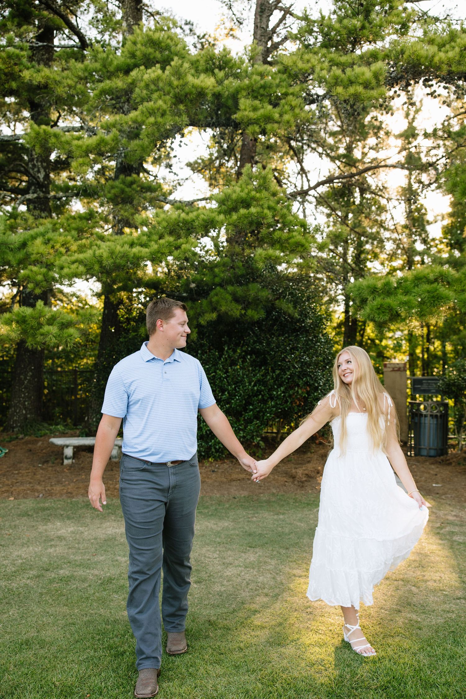 A couple holds hands and smiles while walking on green grass surrounded by tall pine trees in golden hour light.