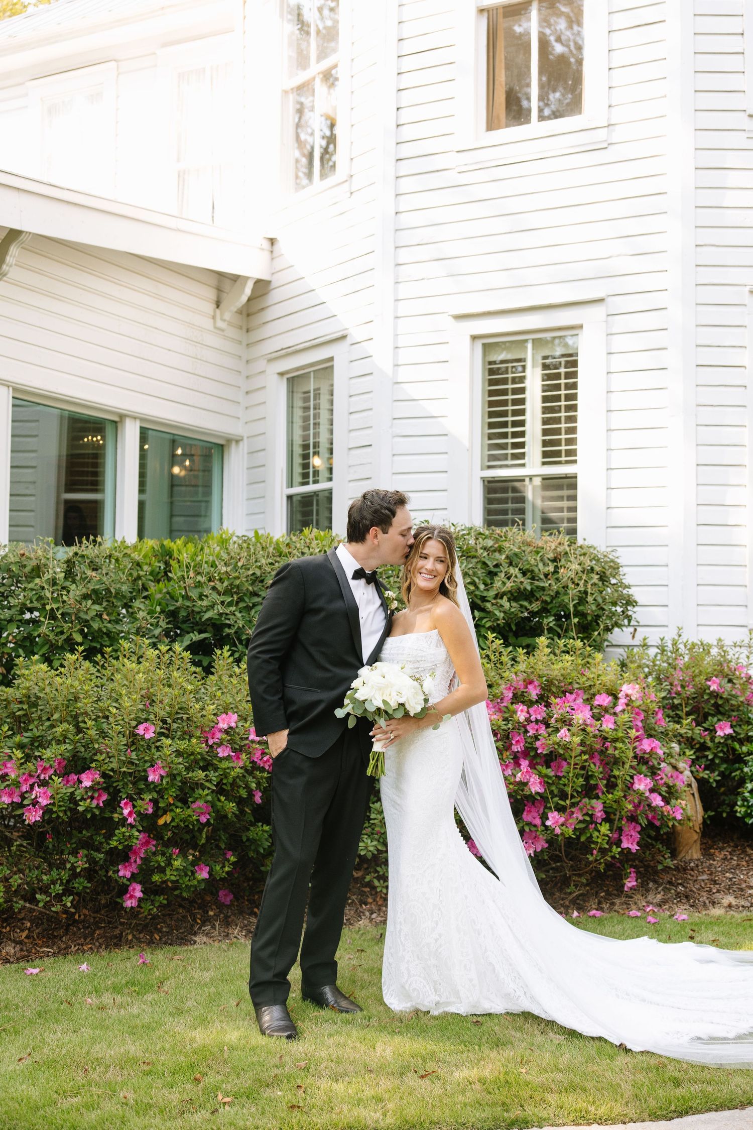 Newlyweds share an intimate moment in front of a white house with blooming pink flowers.