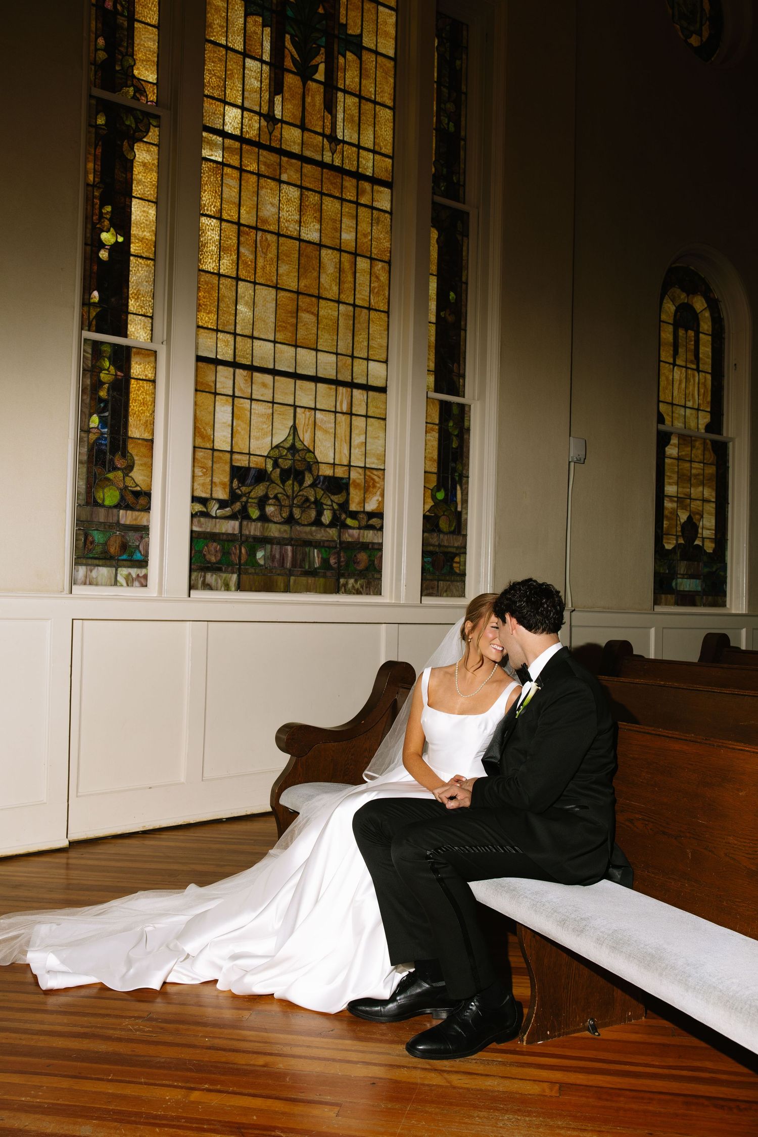 Bride and groom sitting together on a church pew in front of stunning stained glass windows on their wedding day.