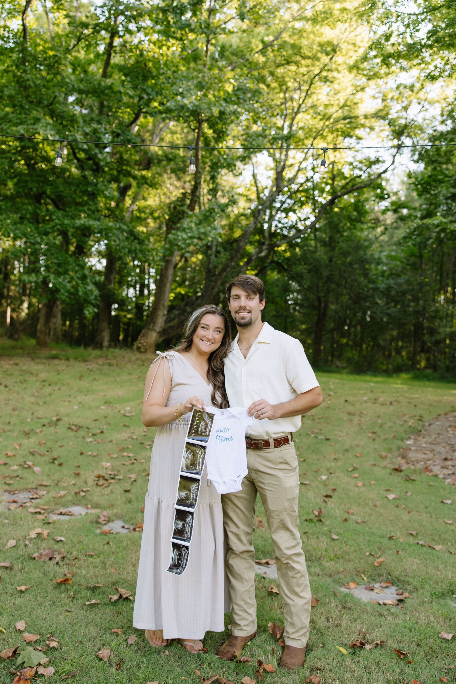 Happy couple stands outdoors holding ultrasound photos and a baby onesie, announcing their pregnancy in a lush green park.