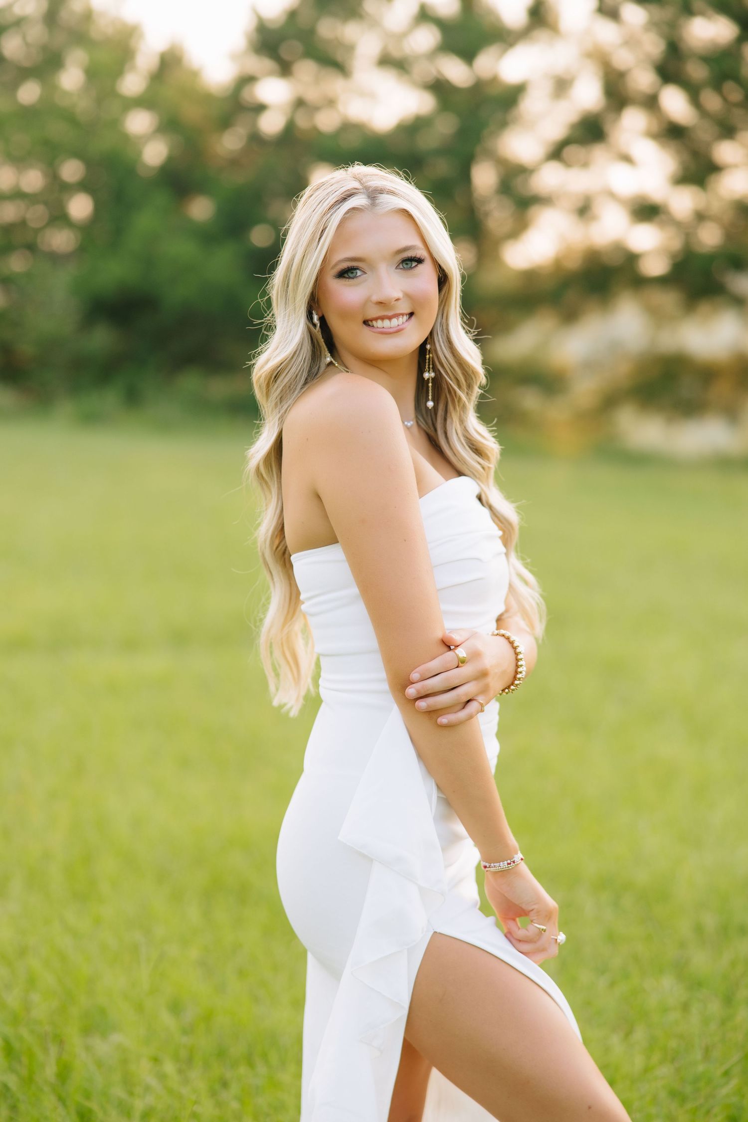 Young woman in white strapless dress posing outdoors on green grass with golden hour sunlight and trees in background.