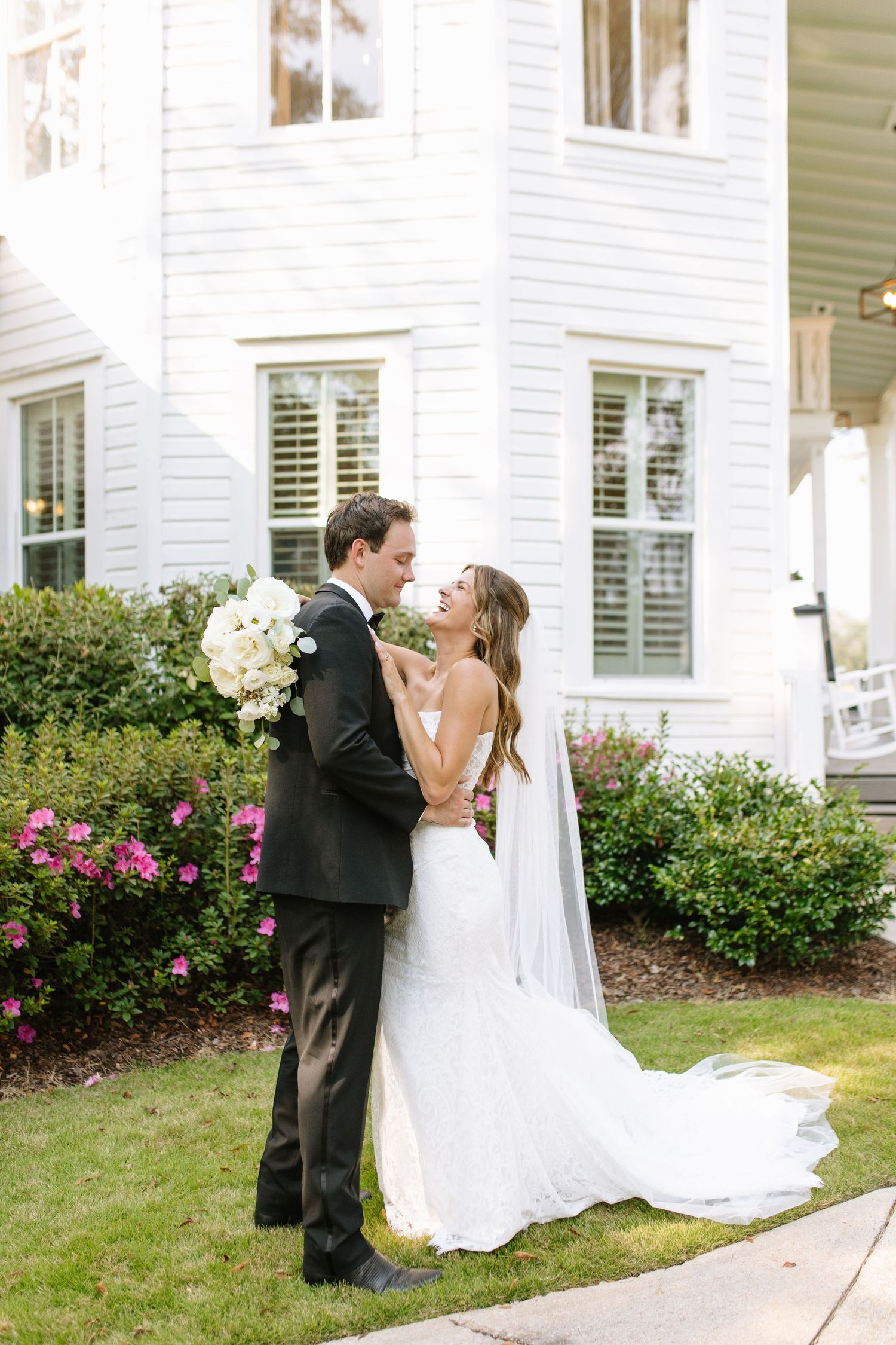 Wedding couple poses by white house with pink flower garden and flowing veil.