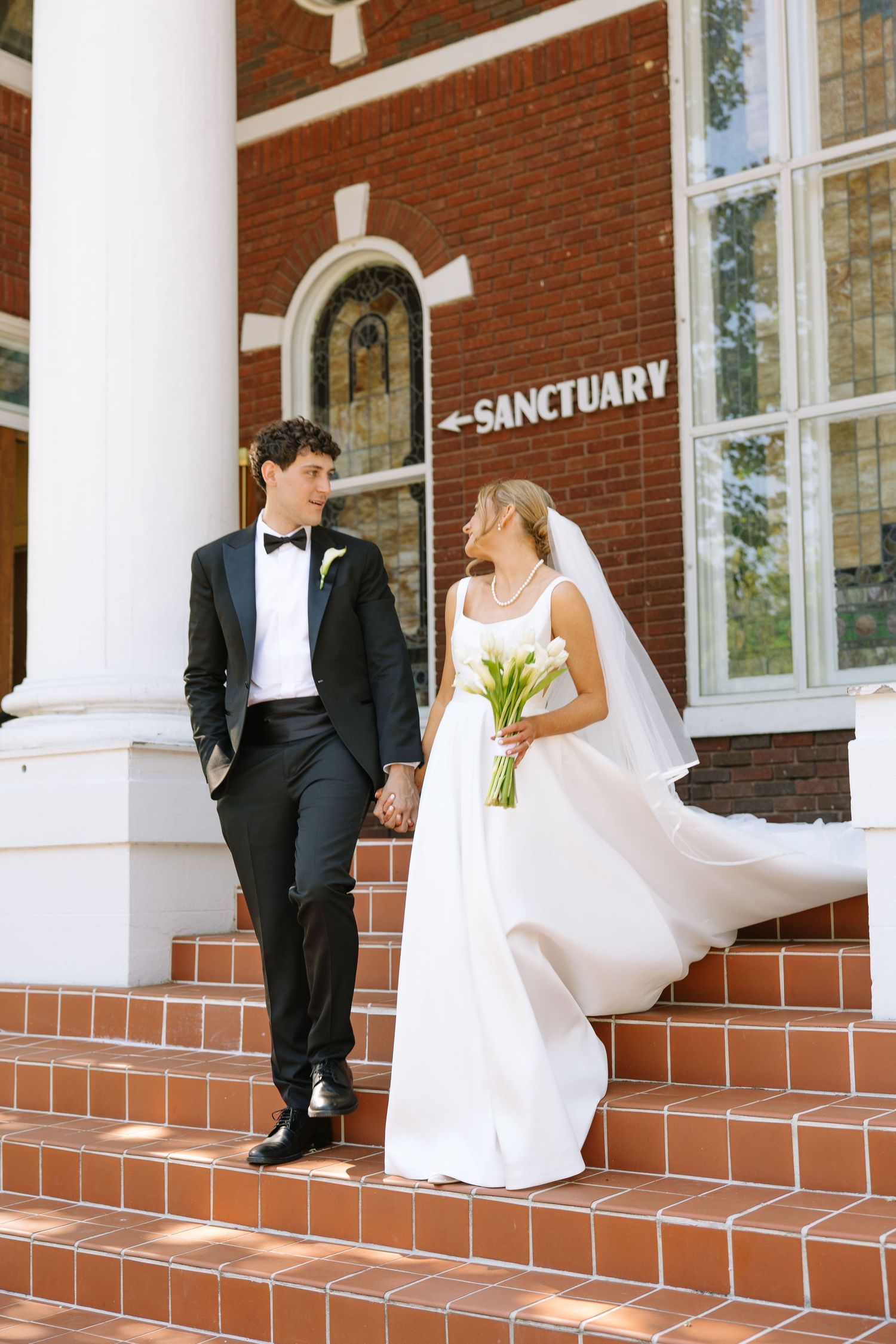 Bride and groom holding hands on church sanctuary steps, she in white gown with veil, he in black tuxedo.