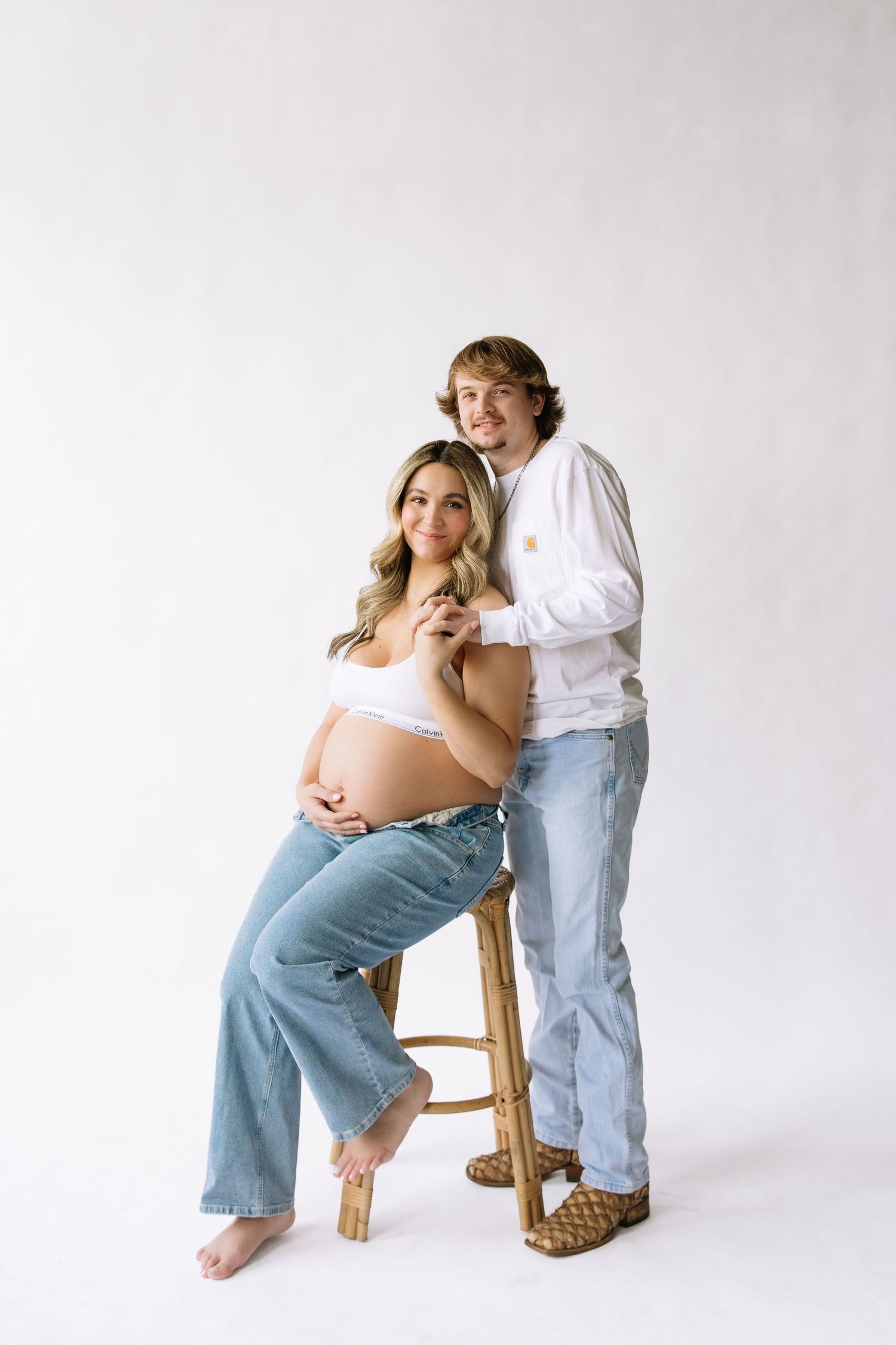 Pregnant woman in white crop top and jeans sits on stool while partner stands behind her in maternity photo.