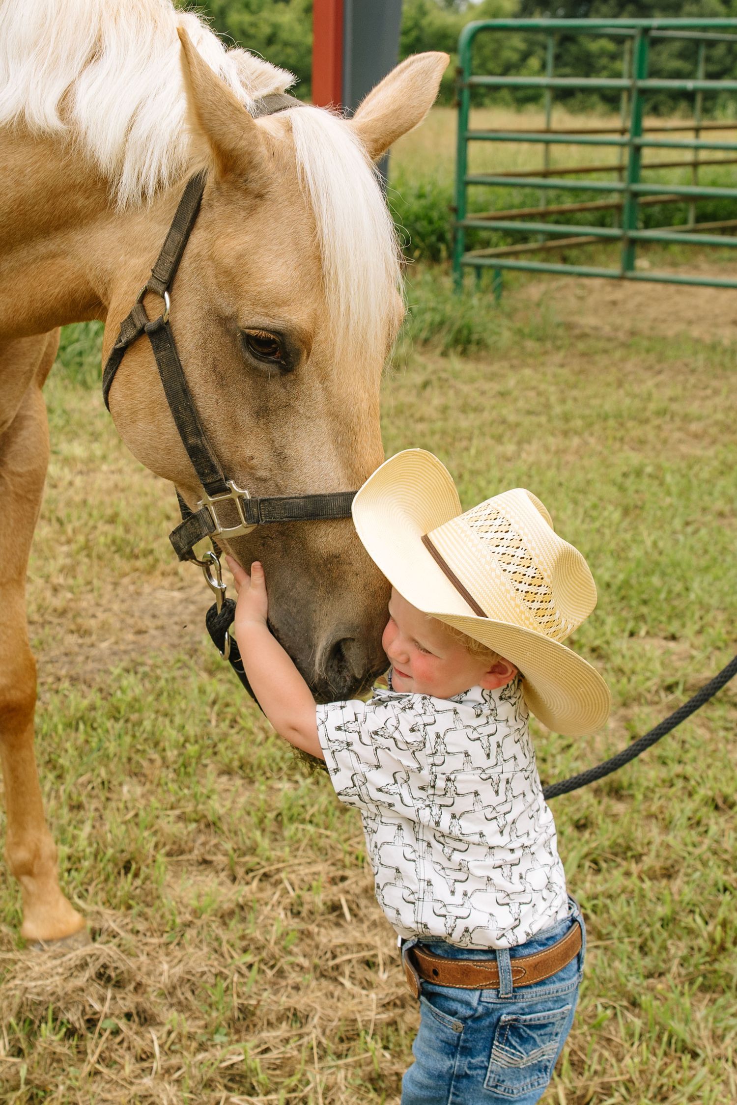 Young child in cowboy hat affectionately hugging a palomino horse near a green fence on a farm.