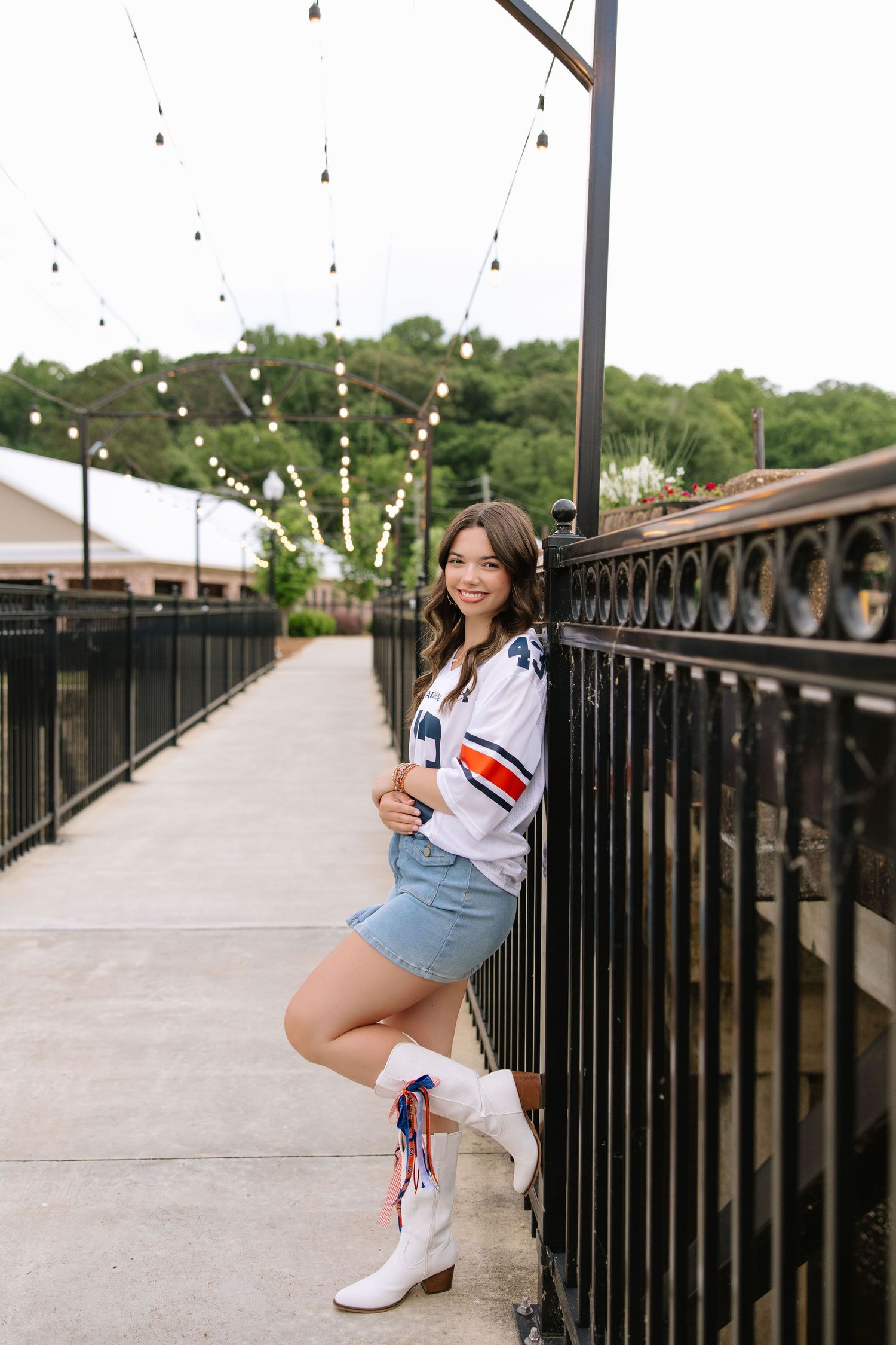 Young woman in white sports jersey, denim skirt, and white cowboy boots poses on a bridge with string lights.