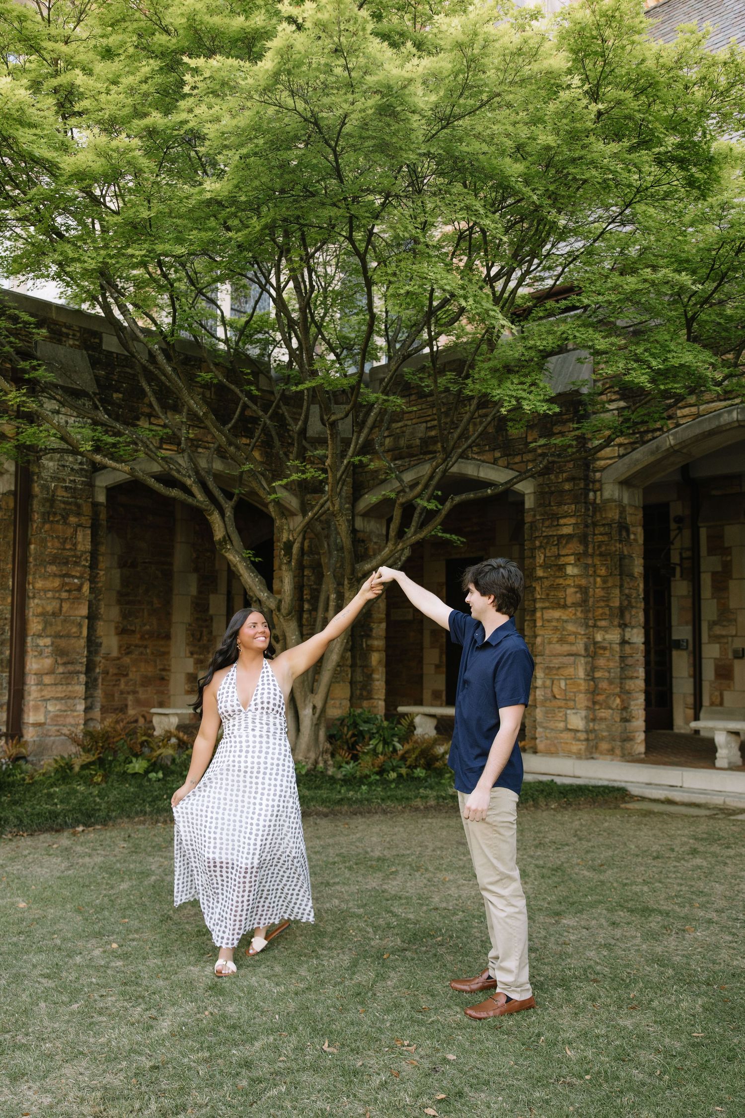 Couple dancing outdoors on green lawn near stone building with lush trees, woman in white dress, man in navy shirt.