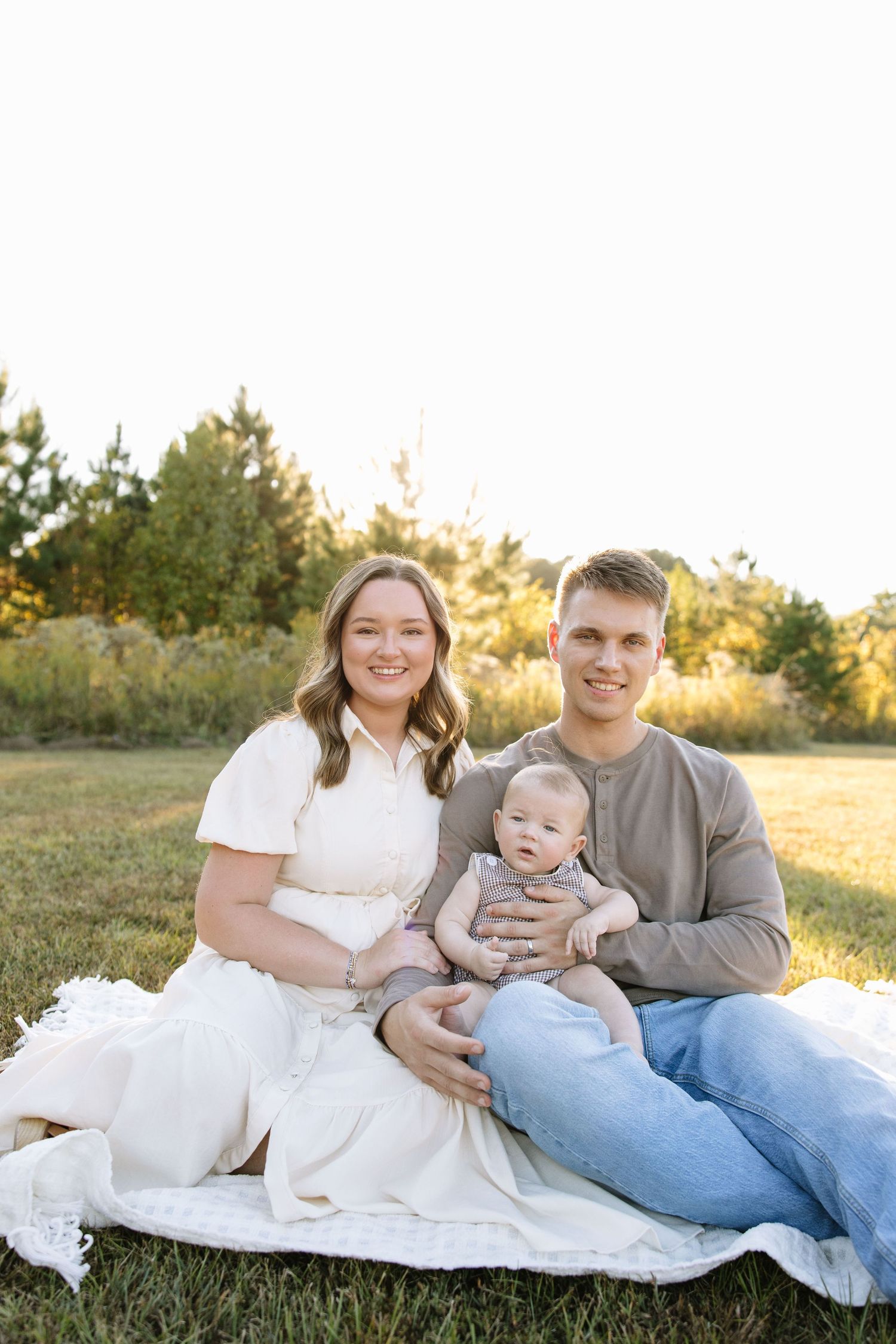 Young family with a baby sitting on a blanket in a sunny green field during golden hour outdoor photo session.
