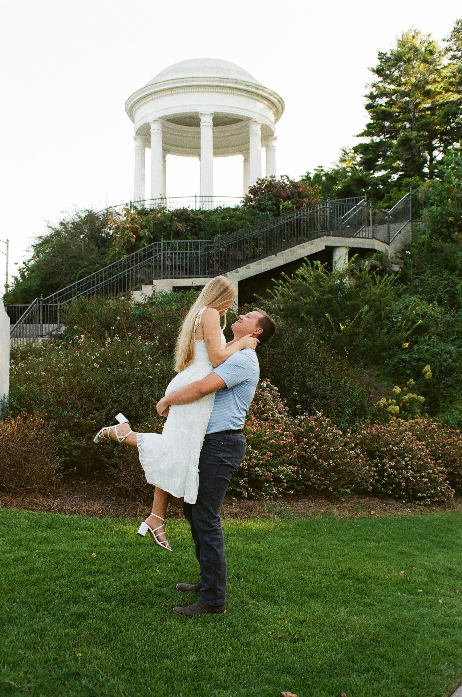 Man lifting woman in white dress, kissing outdoors near white gazebo surrounded by lush greenery on green lawn.