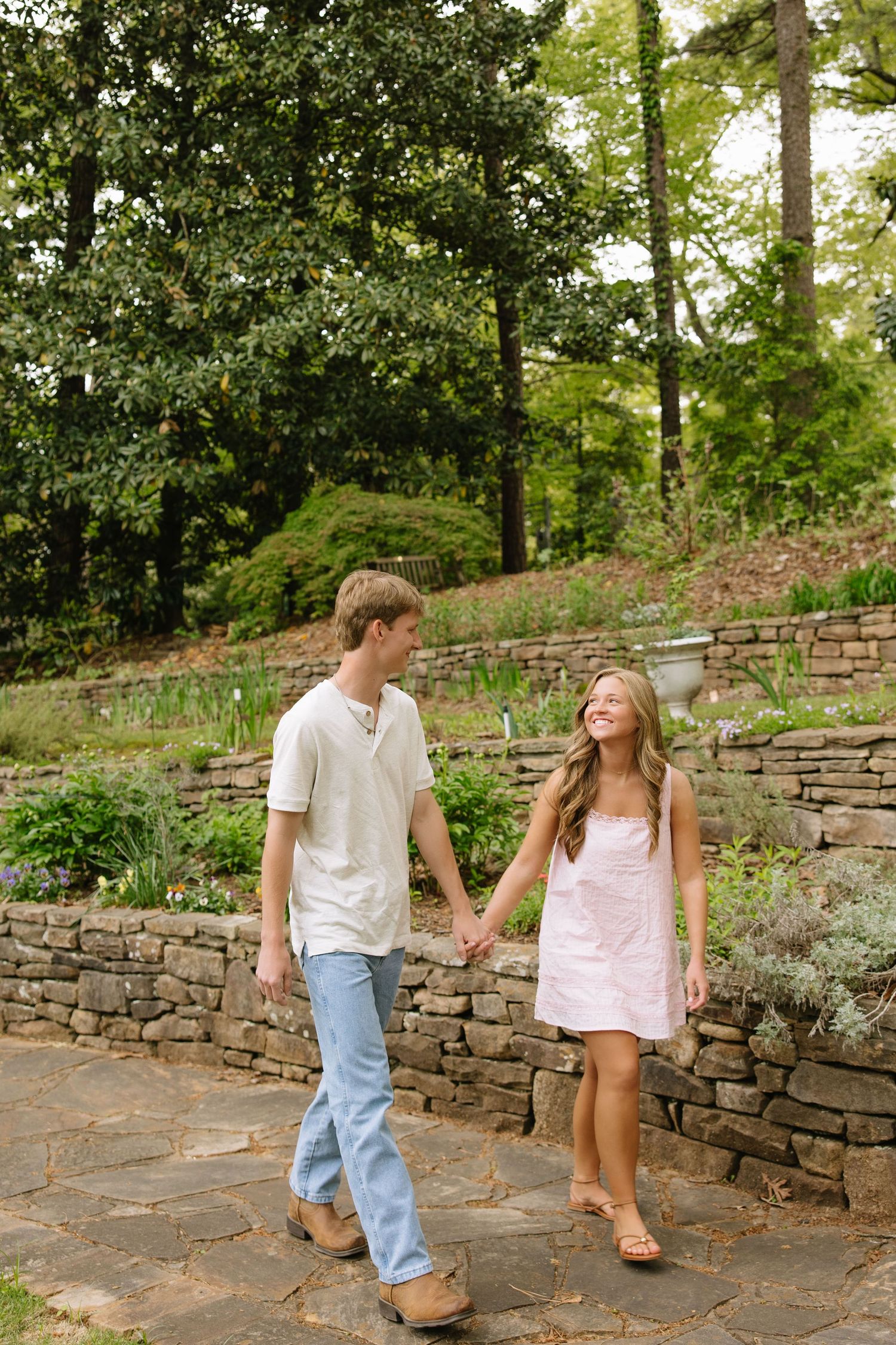 A young couple holds hands and smiles while walking together on a stone path in a lush garden setting.
