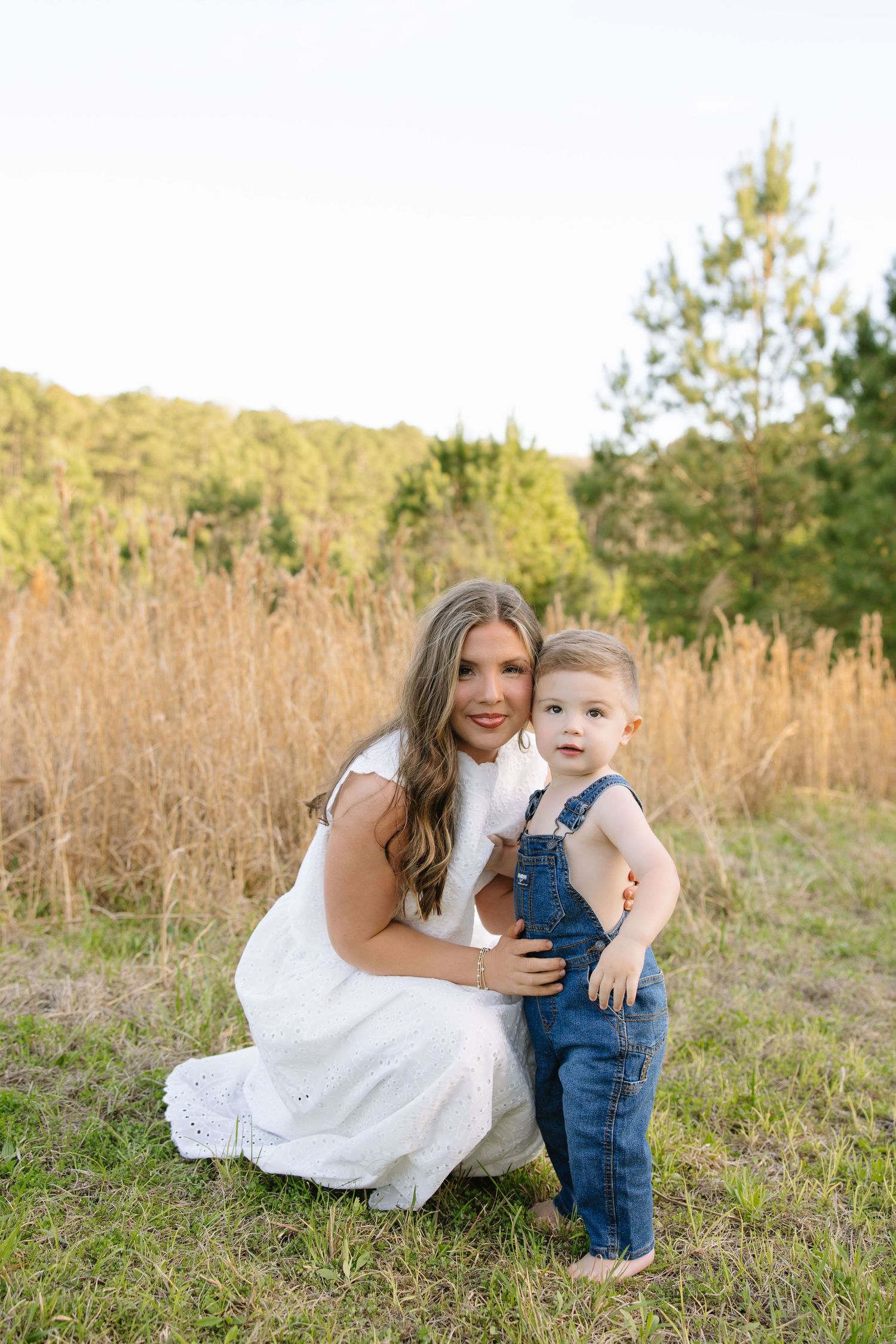 A young woman in a white dress crouches beside a toddler in denim overalls in a sunlit field.