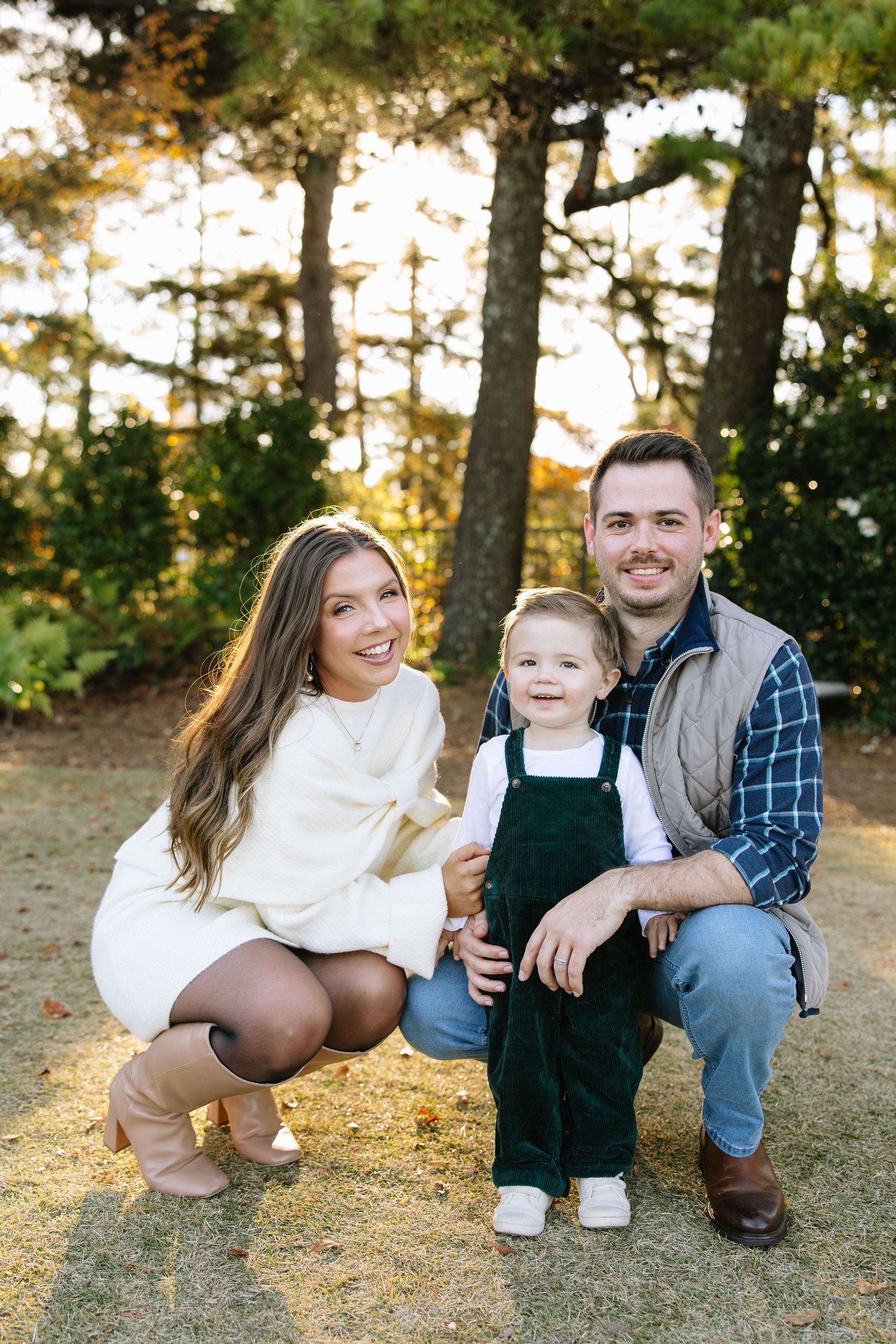 A smiling family of three poses outdoors in fall, with mom, dad, and toddler in green overalls among pine trees.