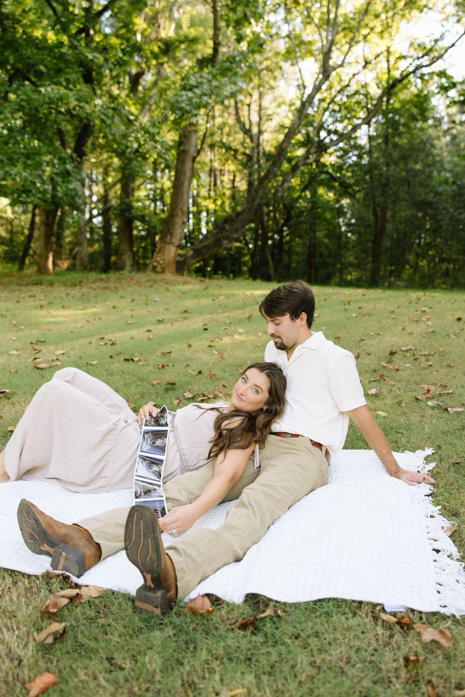 Couple relaxing on white blanket in park, woman holding ultrasound photos, pregnancy announcement outdoor photo.