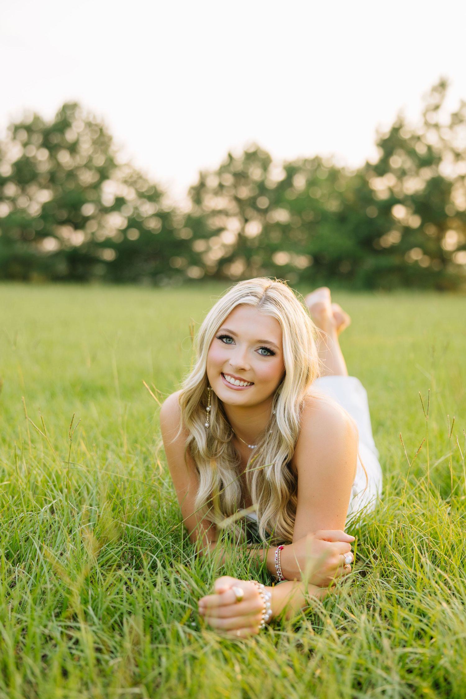 Smiling blonde woman lying in lush green grass field wearing white dress during golden hour outdoor portrait.