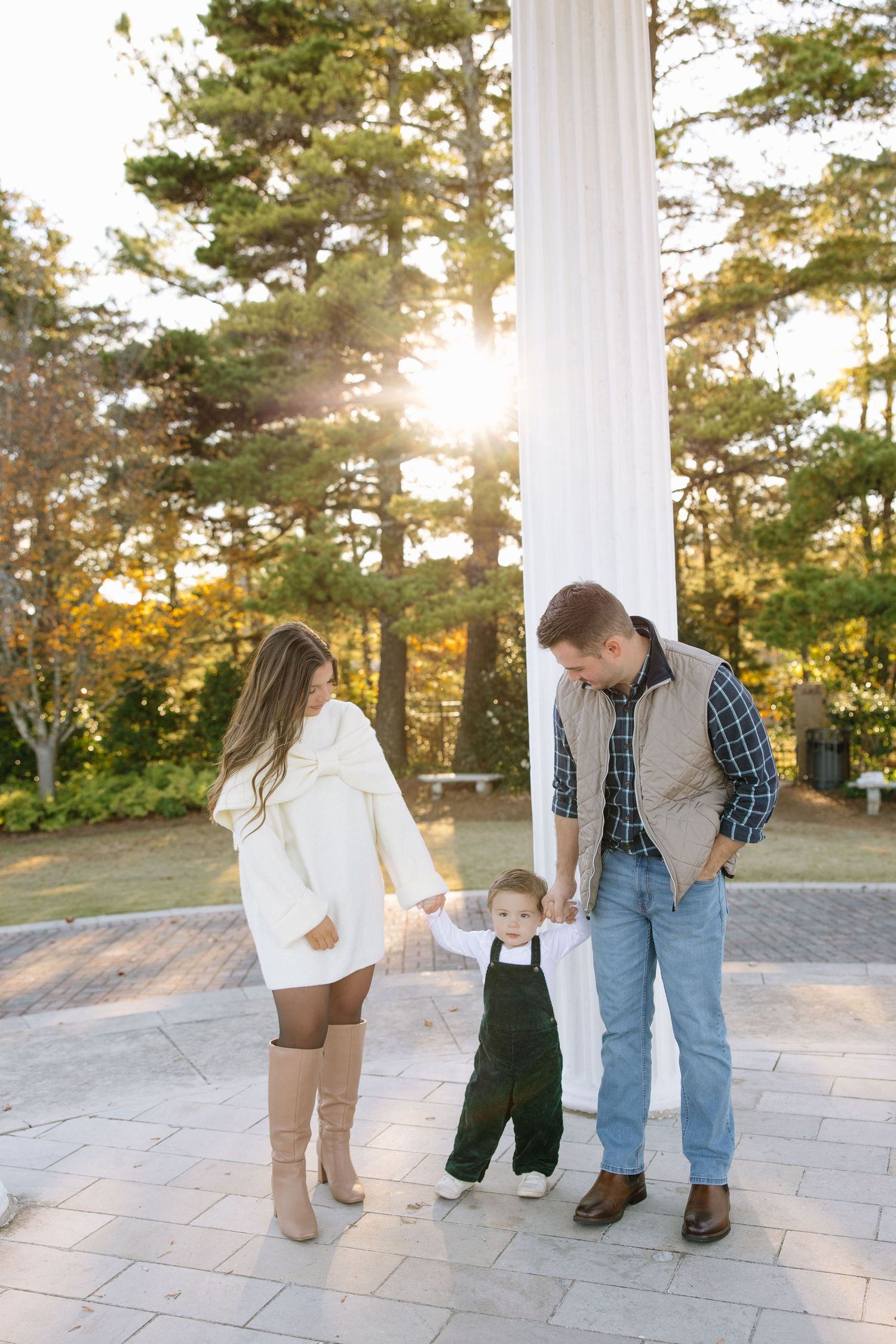 Family of three enjoying a sunny outdoor moment, parents holding toddler's hands near white columns and tall pine trees.