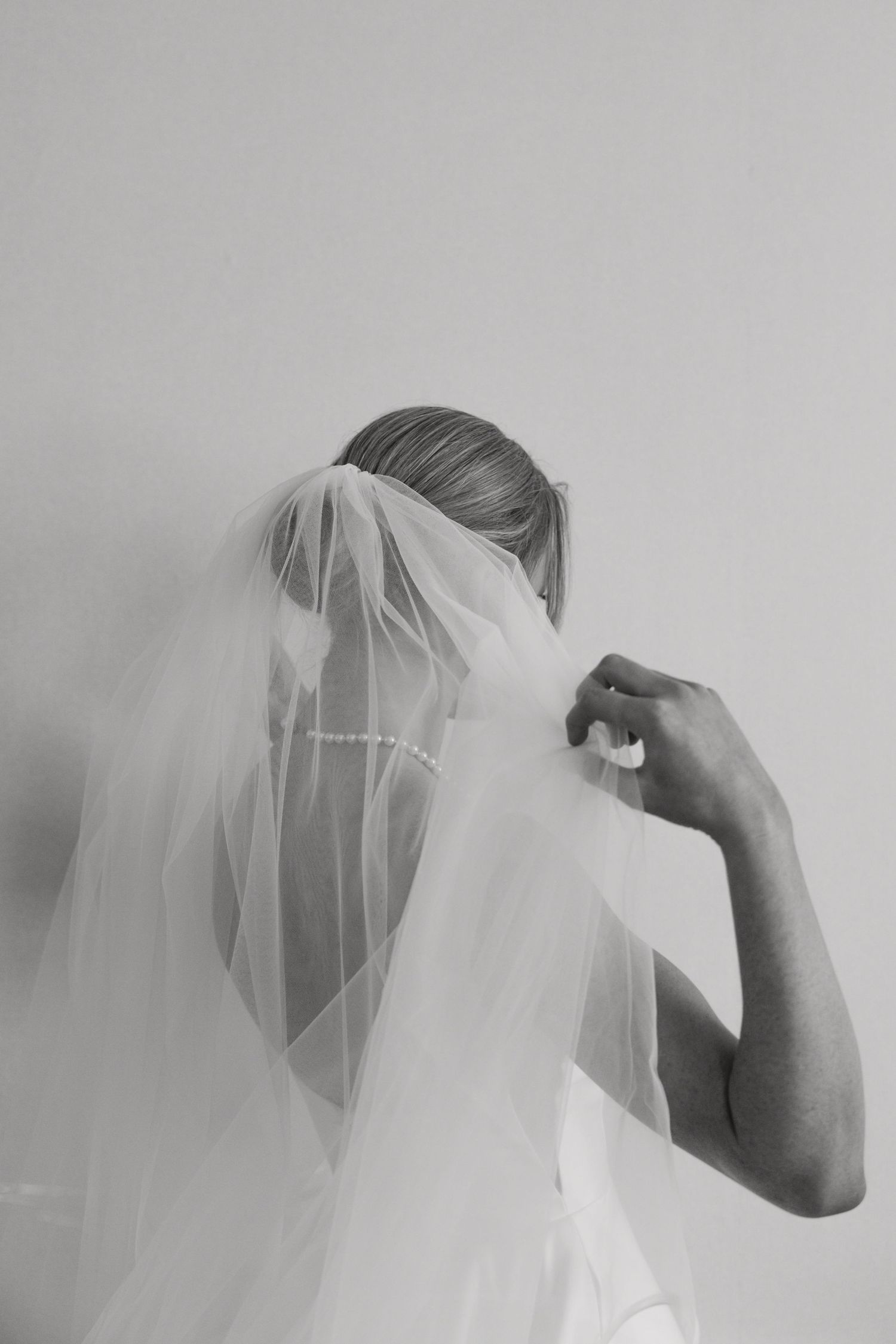 Black and white photo of a bride adjusting her pearl-edged wedding veil from behind.