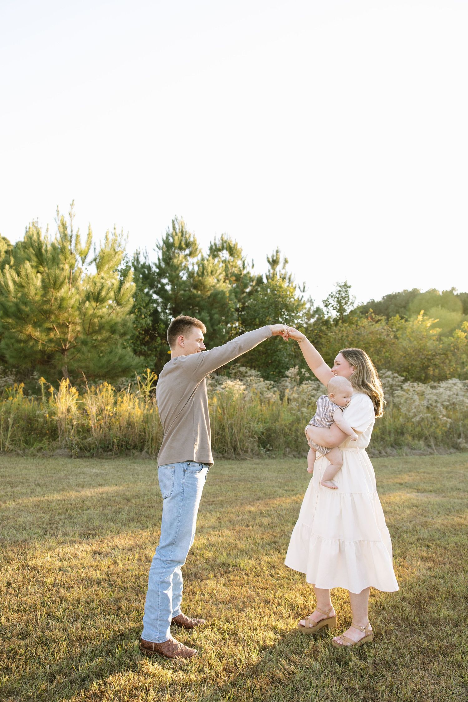 Young couple dancing outdoors in golden hour light, woman in white dress holding baby, man in jeans, green trees behind.